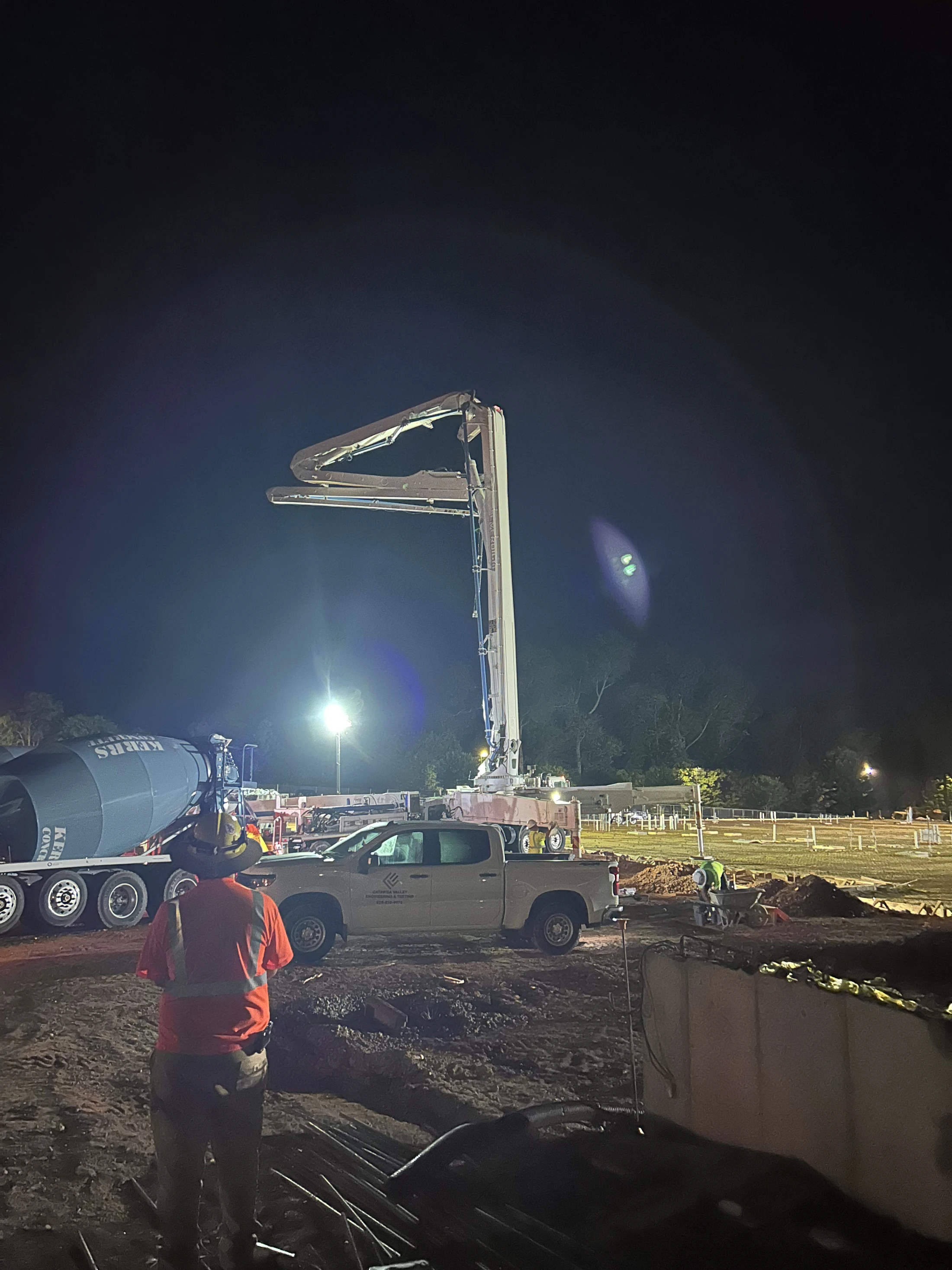 A nighttime construction site with a concrete mixer and a large pump truck with its boom extended. Workers in safety gear are present under bright lights.