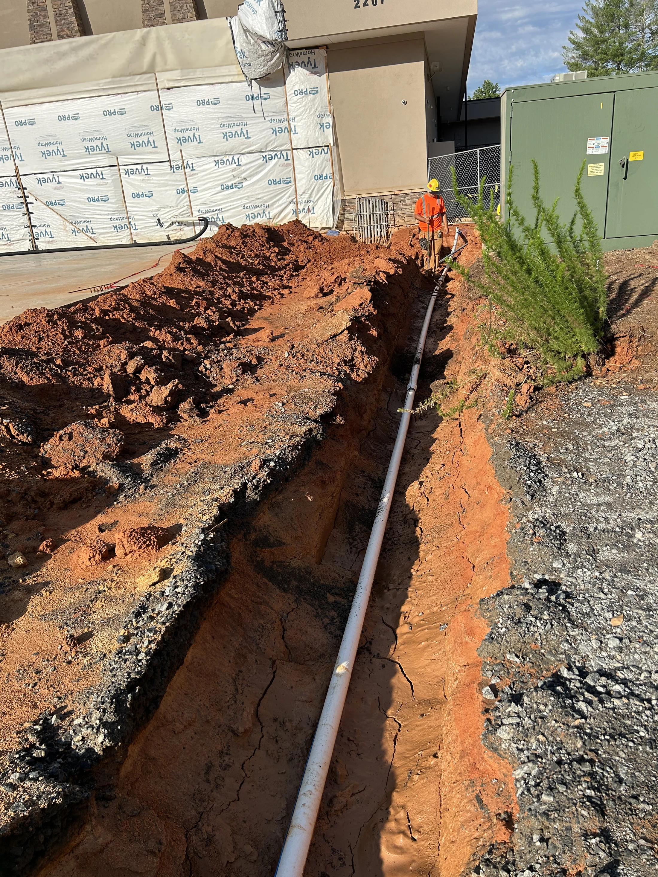 A construction worker in a safety vest stands in a long, deep trench laying a white pipe next to a building under construction.
