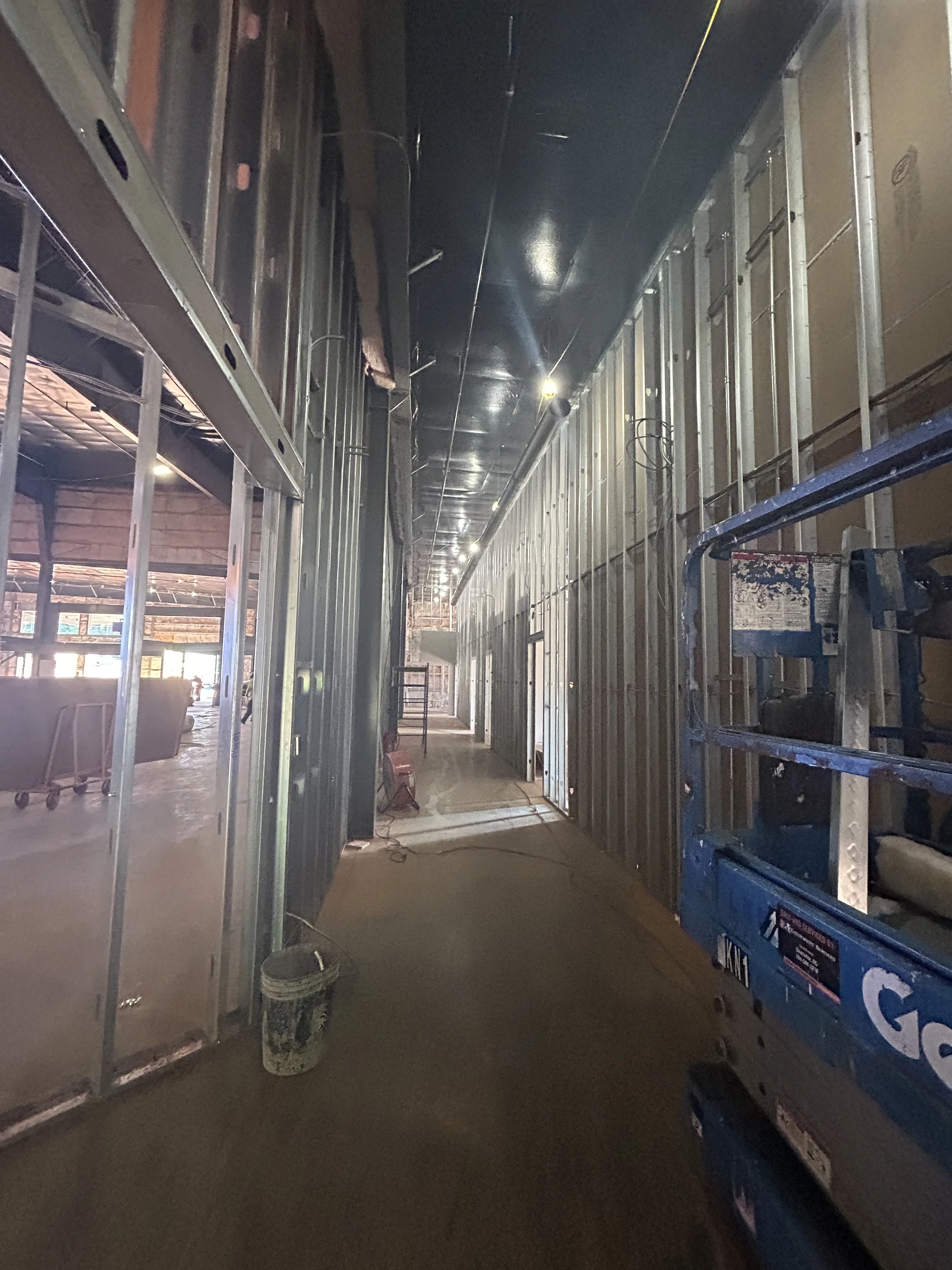A long hallway in a building under construction, showing exposed metal wall studs, a dark ceiling, concrete floors, and a blue aerial lift on the right.