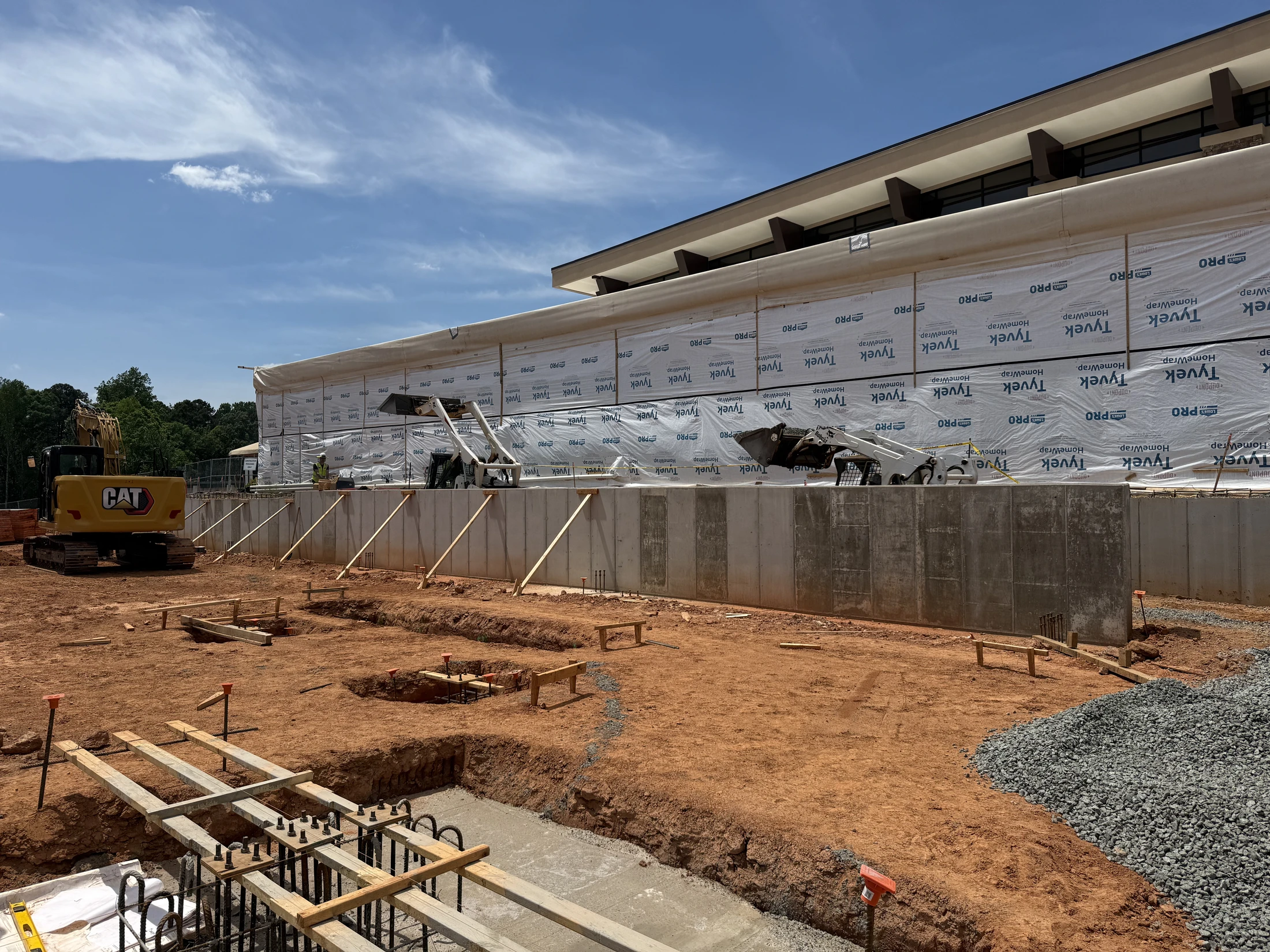 A wide shot of a construction site with a new building foundation. The foreground shows trenches, rebar, and formwork in red dirt under a blue sky.