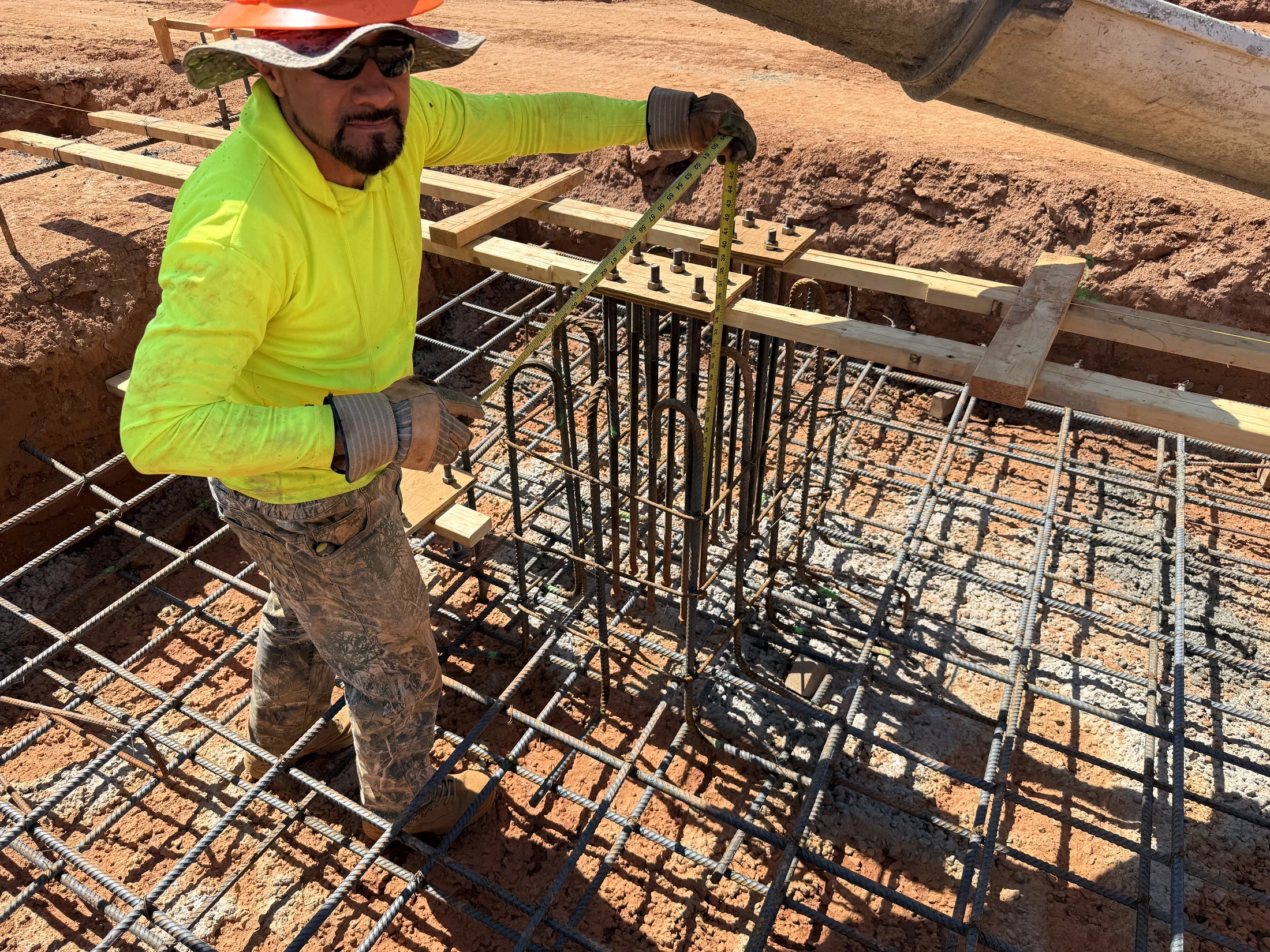 A construction worker in a hard hat and safety vest measures a steel rebar cage with a tape measure before pouring a concrete foundation.