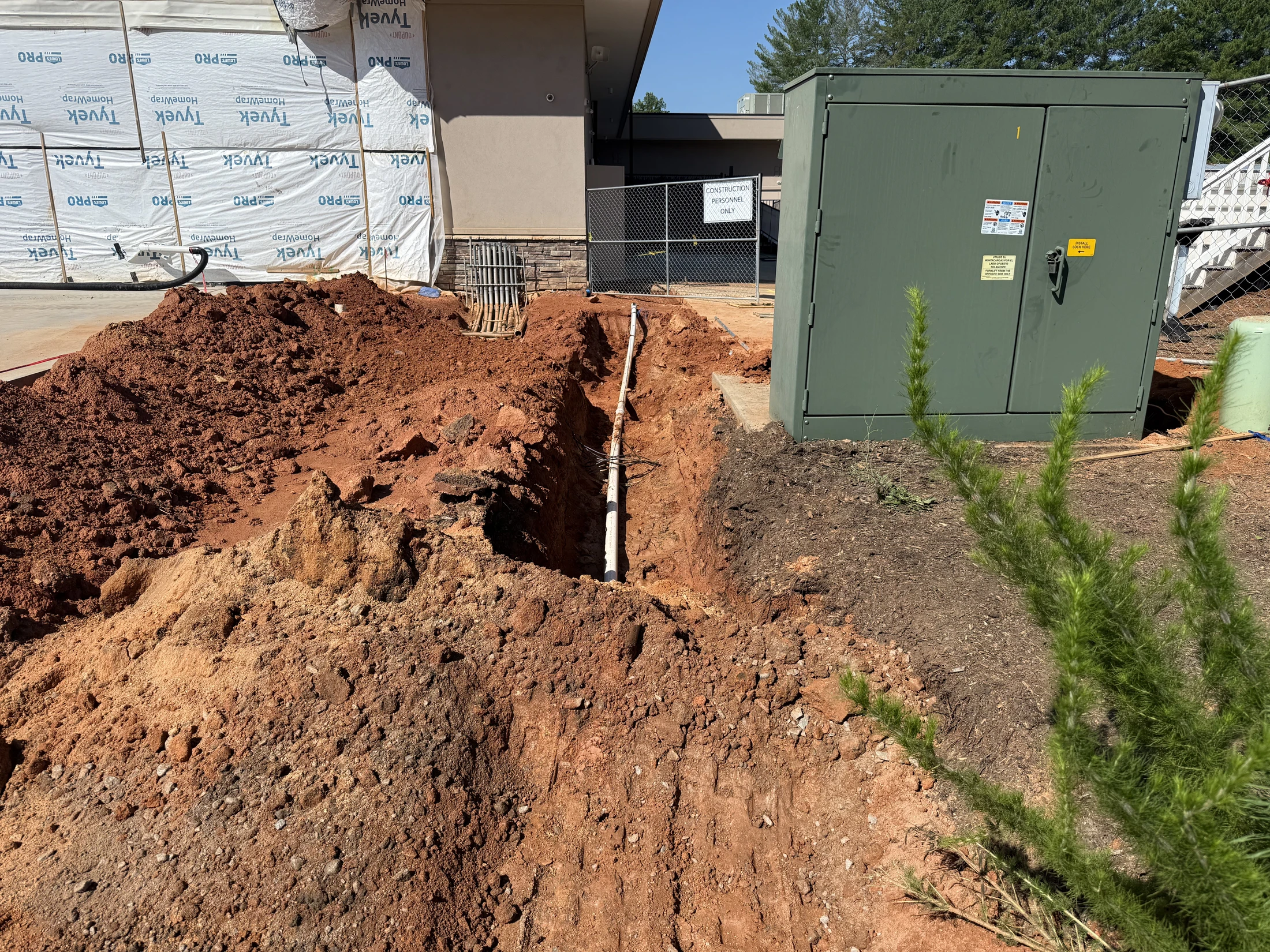 A construction site with a deep trench dug in red clay soil, containing a white pipe that leads to a large green electrical box next to a new building.