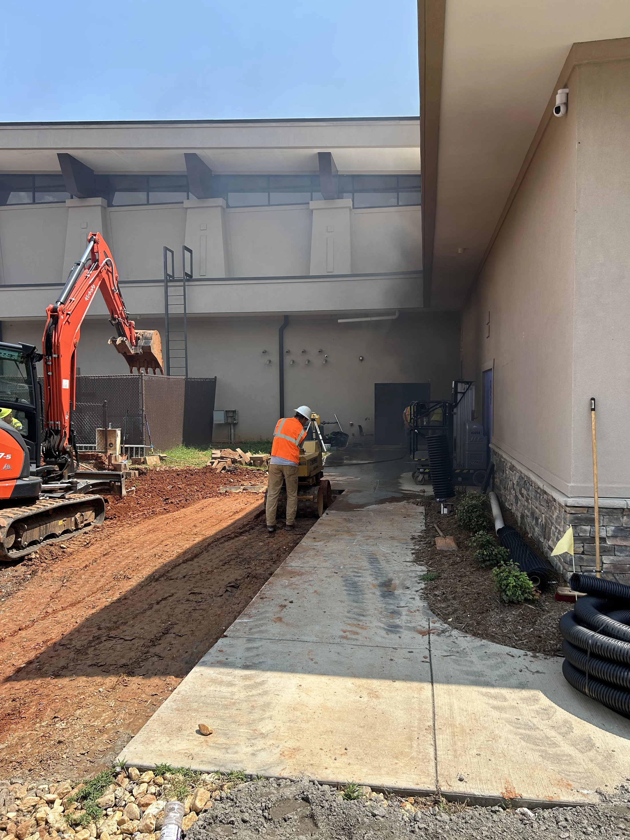A construction worker in an orange safety vest operates a machine on a sidewalk, with an orange excavator on the dirt nearby, next to a building.