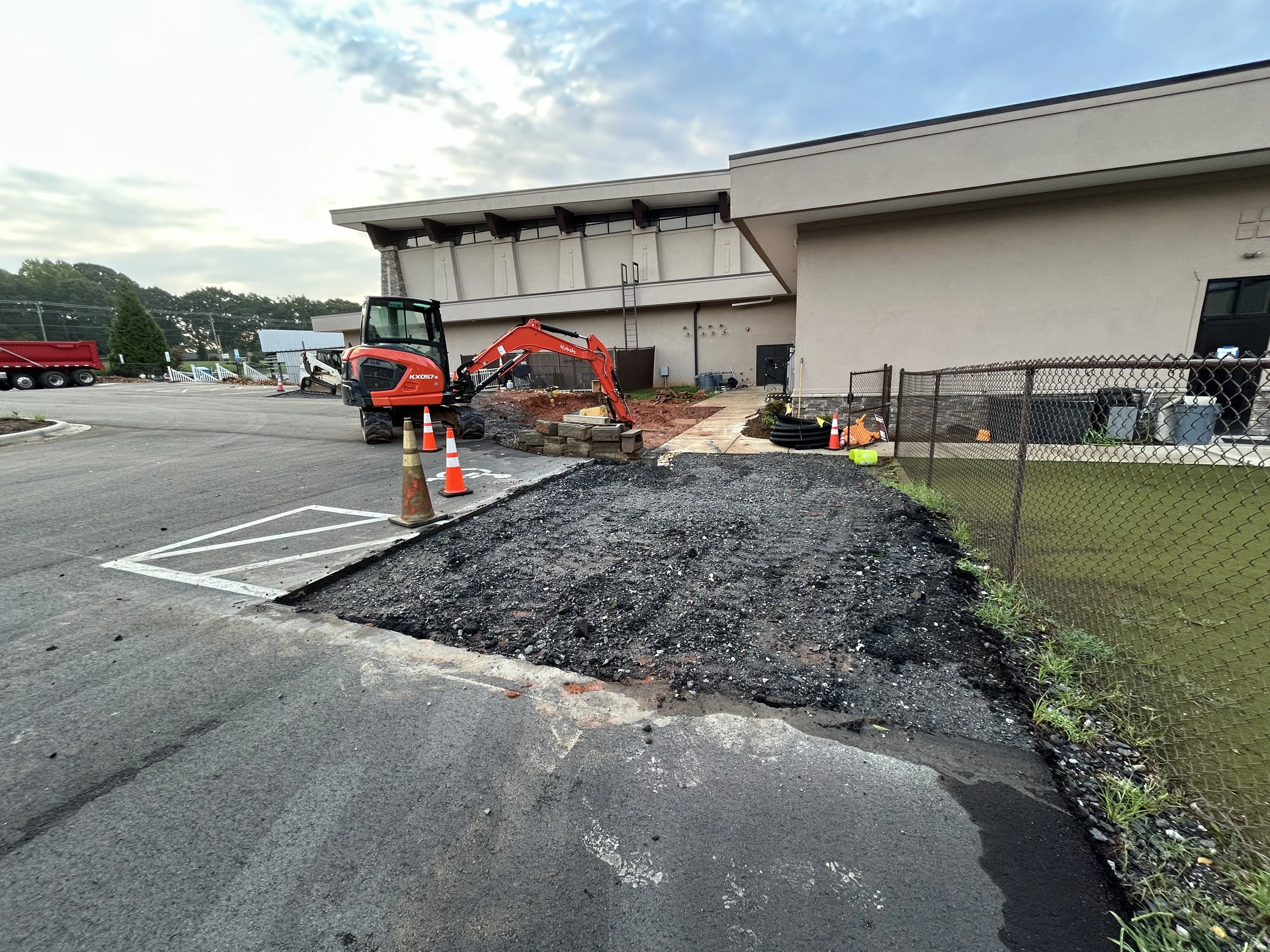 An orange mini excavator at a construction site where a section of an asphalt parking lot has been removed, with traffic cones and a building in the background.