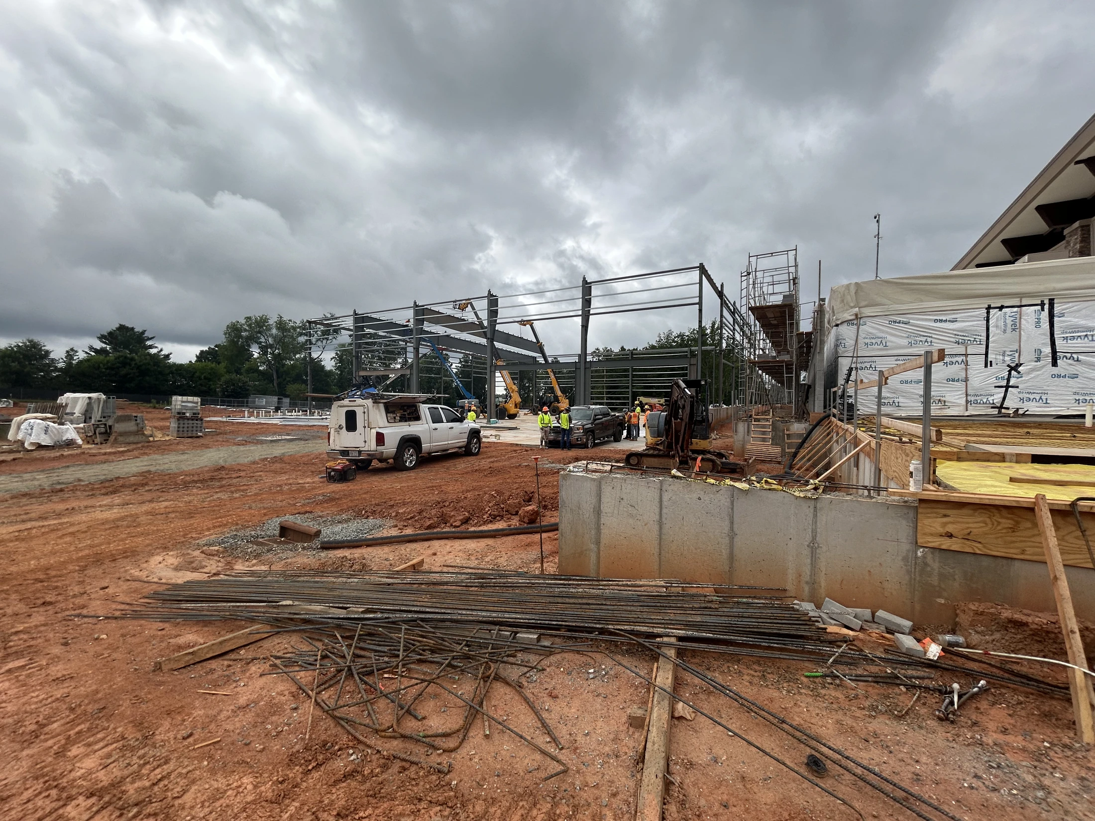 A wide view of a construction site on a cloudy day. A steel building frame is being erected in the background, with rebar and machinery in the dirt foreground.