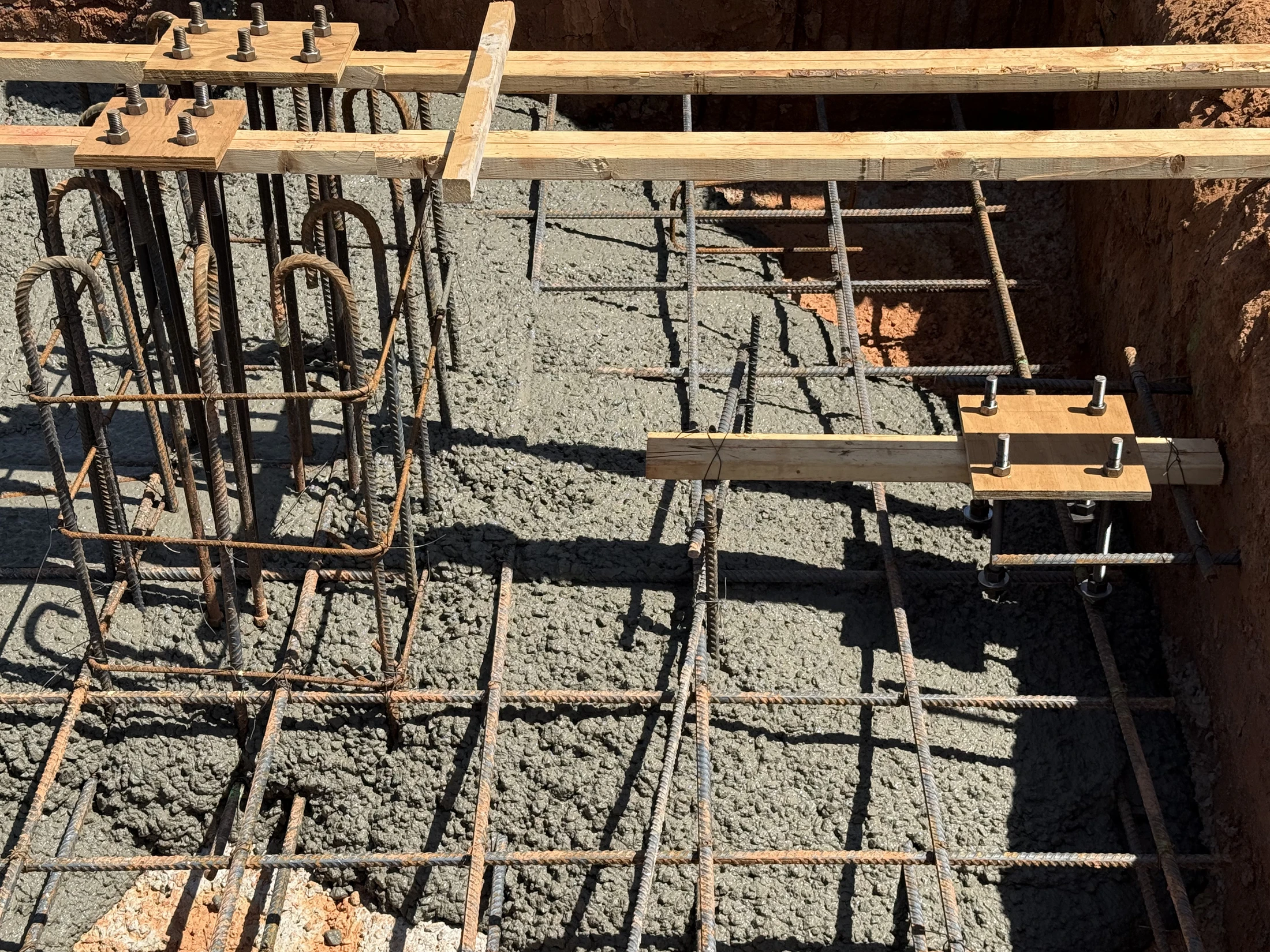 A close-up of a building foundation under construction. Wet concrete is poured around a grid of steel rebar, with anchor bolts set in place by wooden forms.