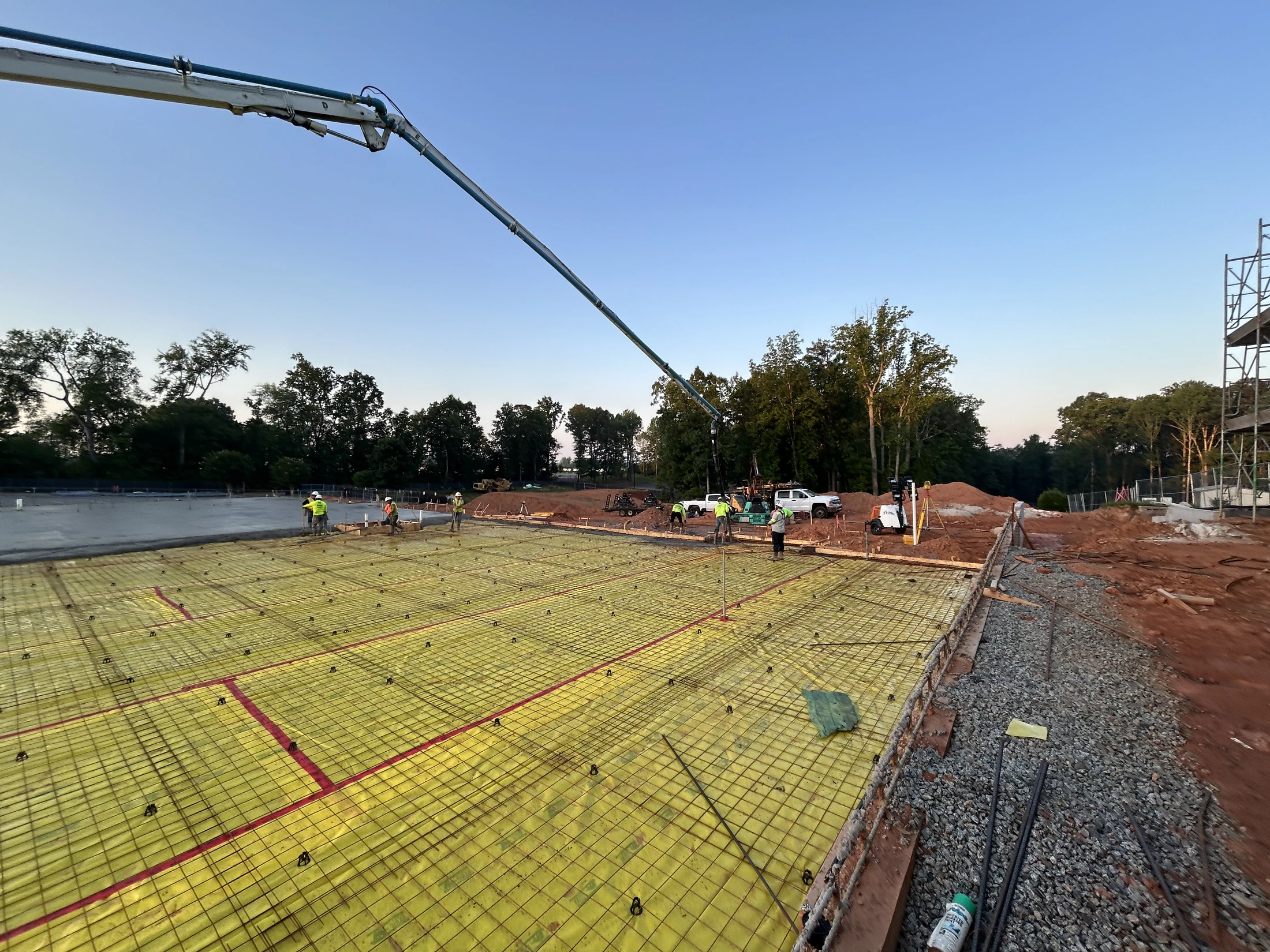 A wide-angle view of a construction site at dawn, with workers preparing a large foundation with a yellow barrier and rebar, a concrete pump boom overhead.