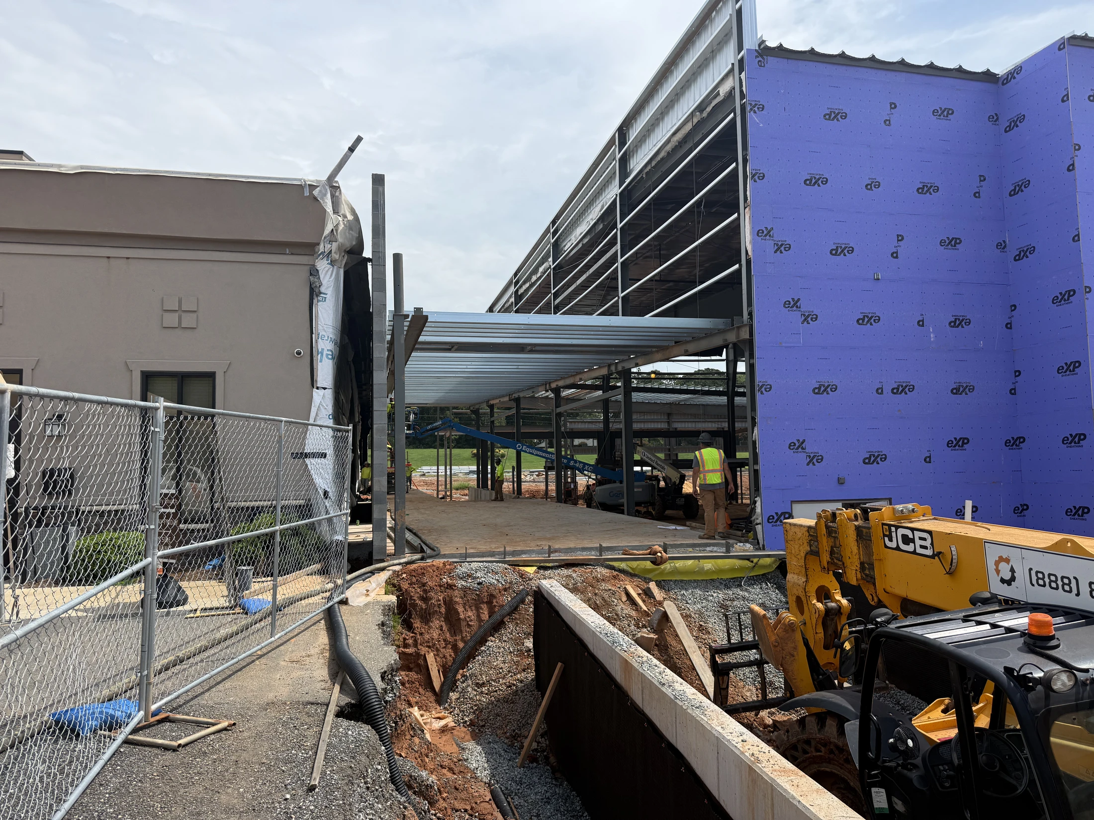 A construction site showing a new steel-framed building with purple exterior sheathing being built next to an existing tan building, with a worker on site.