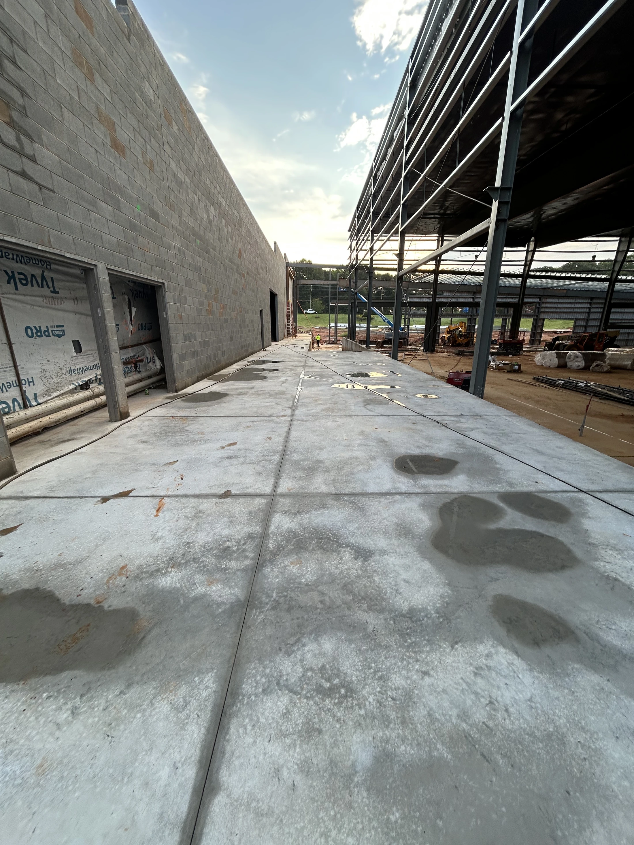 A wide-angle view down a wet concrete walkway between a cinder block wall and the steel frame of a building under construction at dusk.