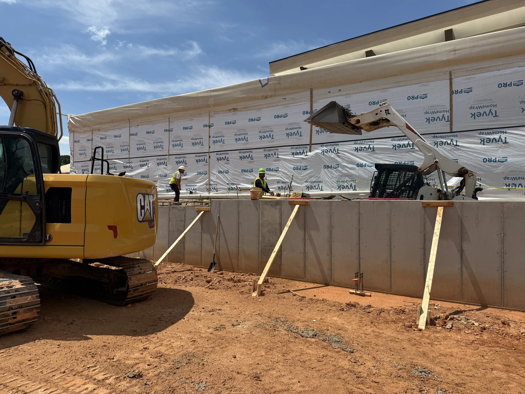 A construction site with a yellow excavator and white skid-steer next to a concrete foundation where two workers in hard hats are standing.