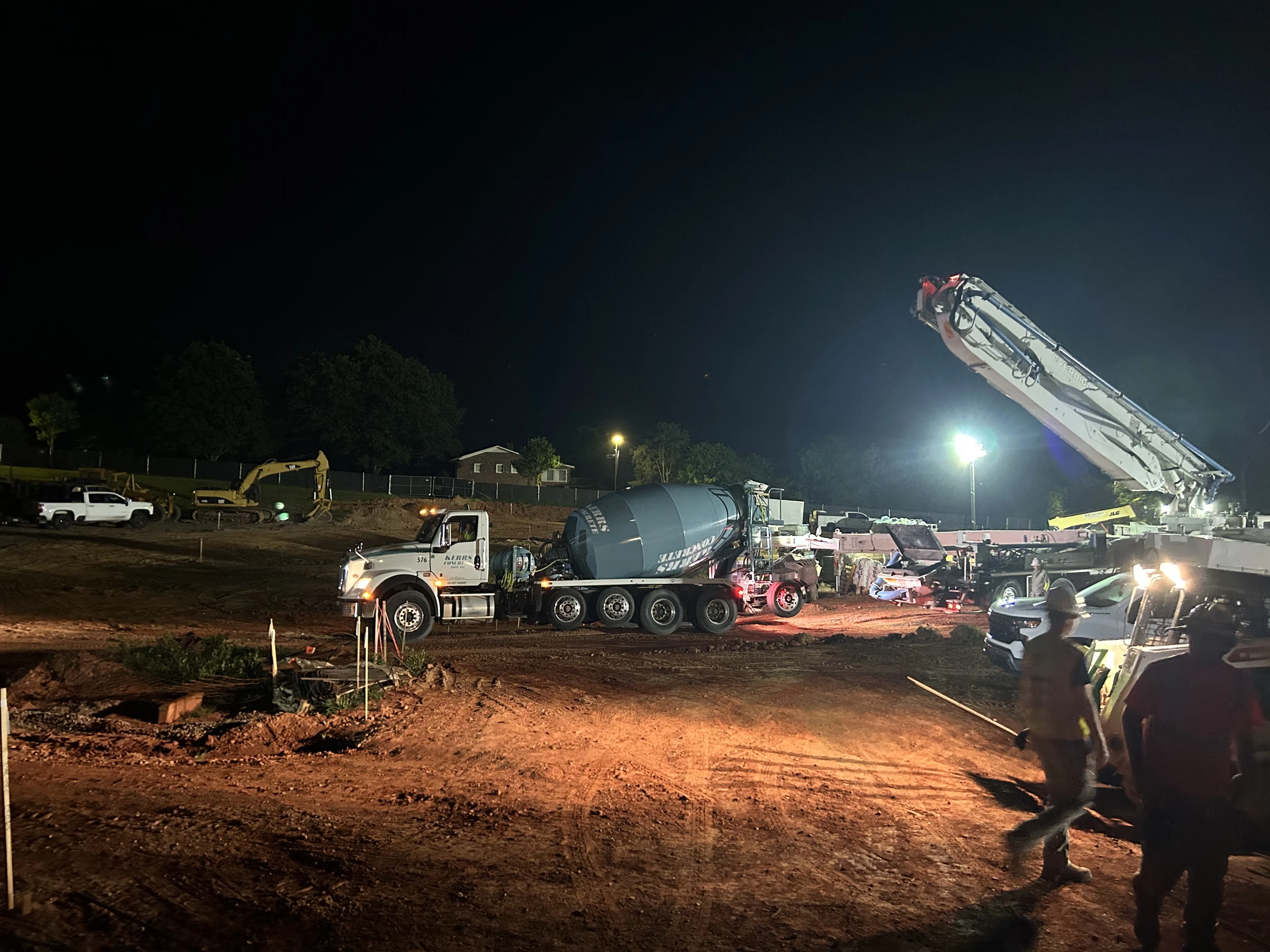 A busy construction site at night, illuminated by bright lights. A concrete mixer truck and a concrete pump are at work with construction crew nearby.