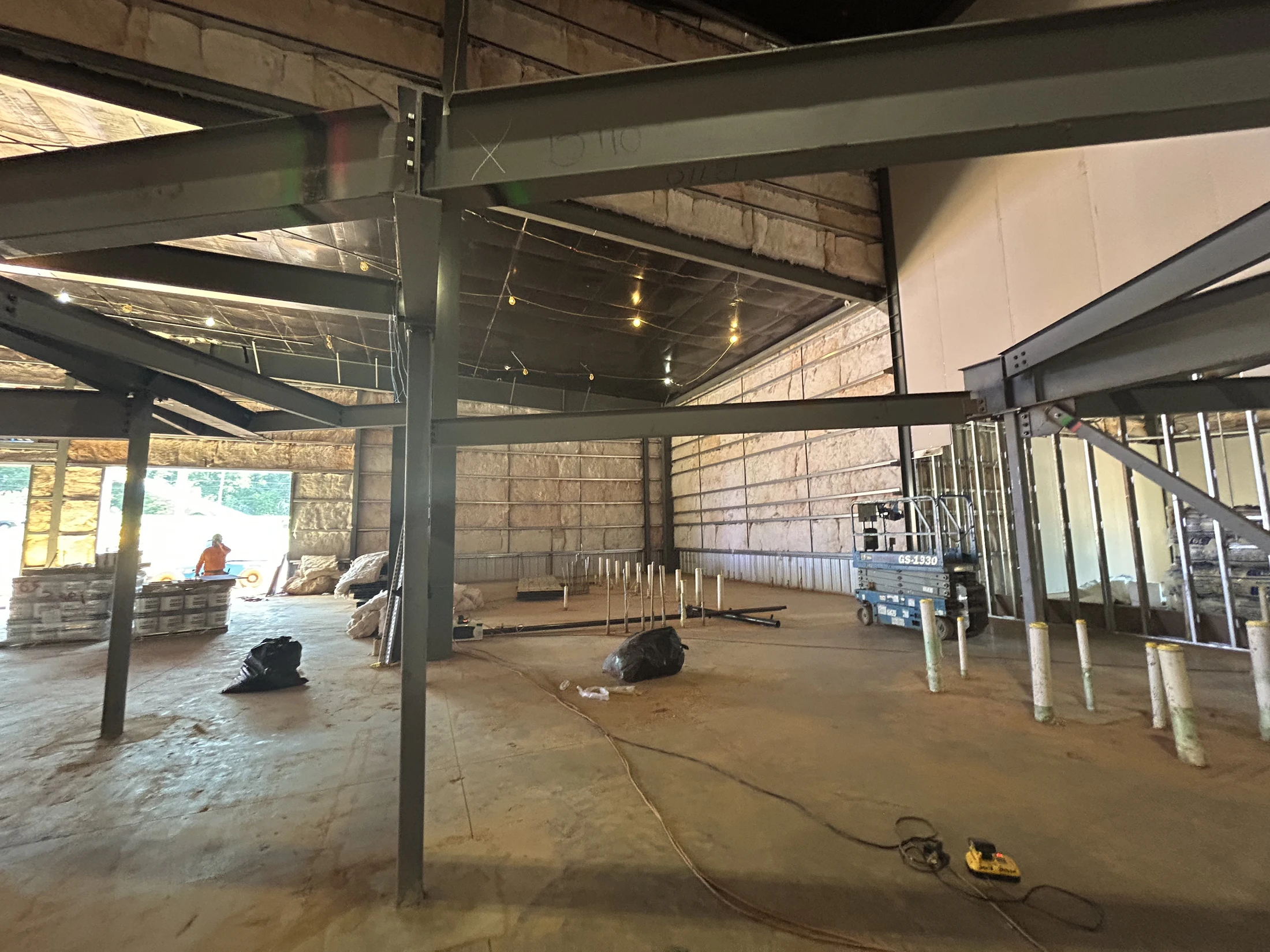 The unfinished interior of a large building with steel support beams, exposed insulation on the walls, and a concrete floor with pipes and equipment.