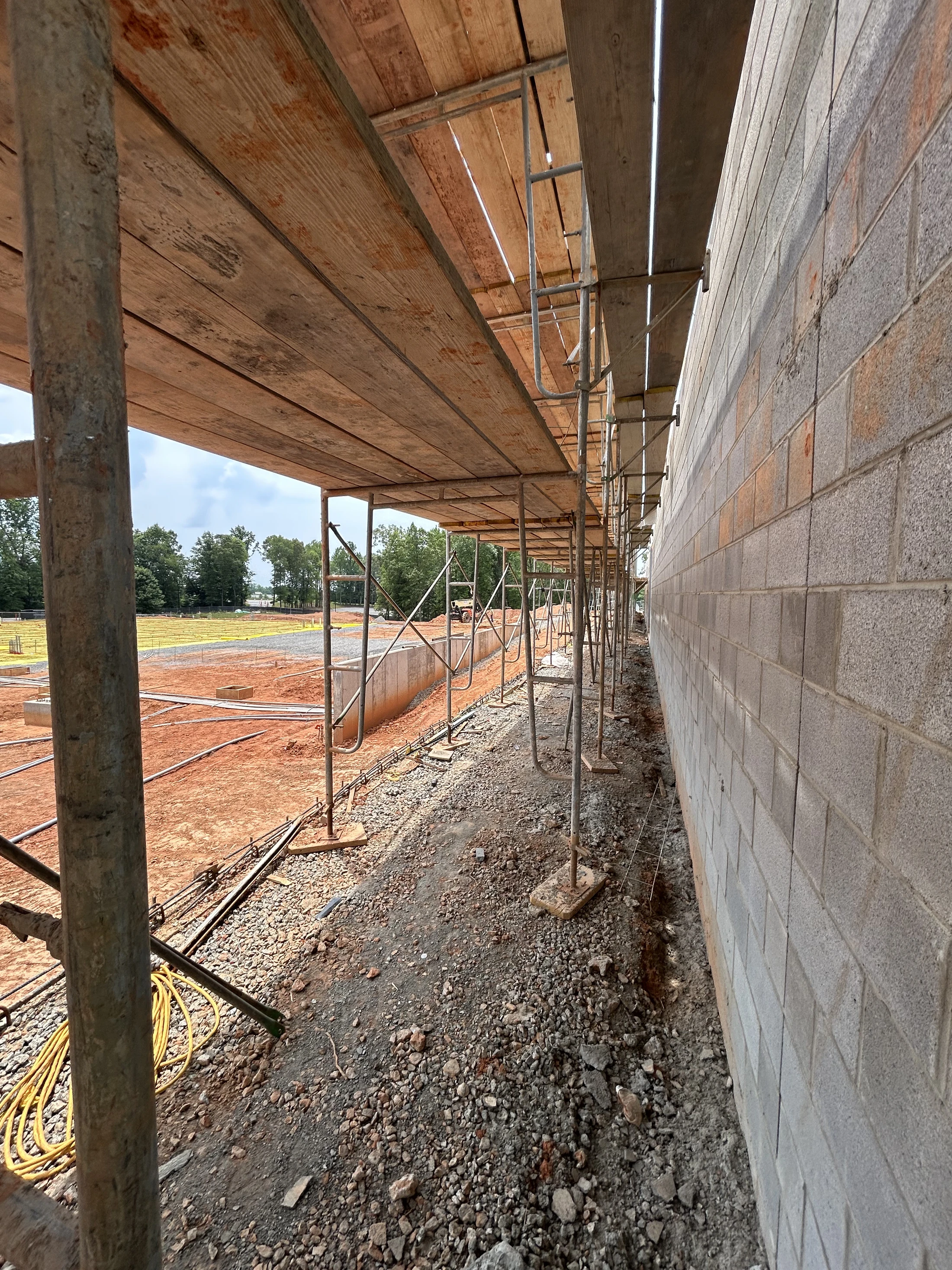 A ground-level view looking down a long line of scaffolding next to a grey cinder block wall at a construction site with red dirt and gravel.