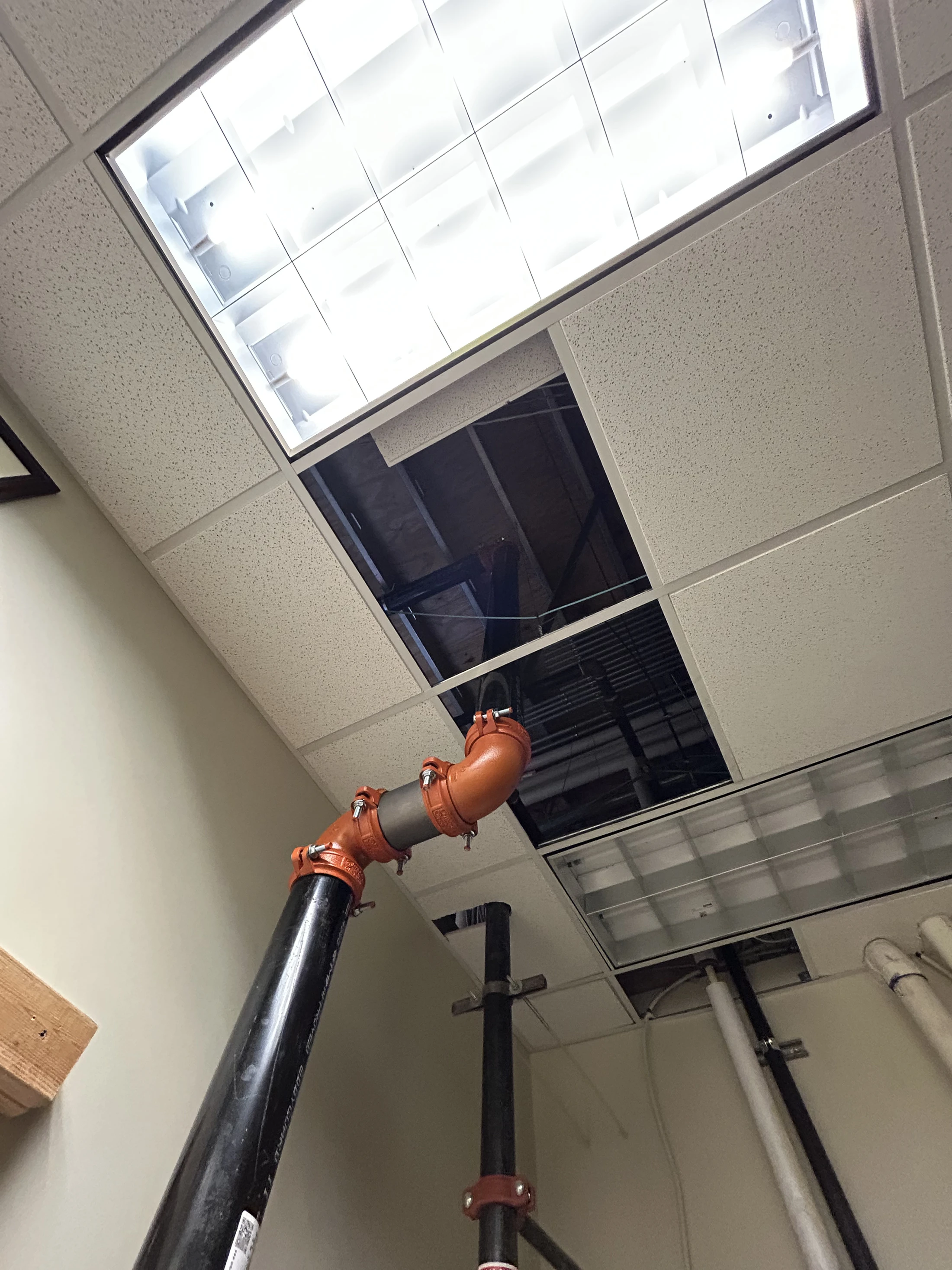 A low-angle shot of an office ceiling with missing tiles, revealing the dark space above. A large black pipe with orange fittings extends up into the opening.