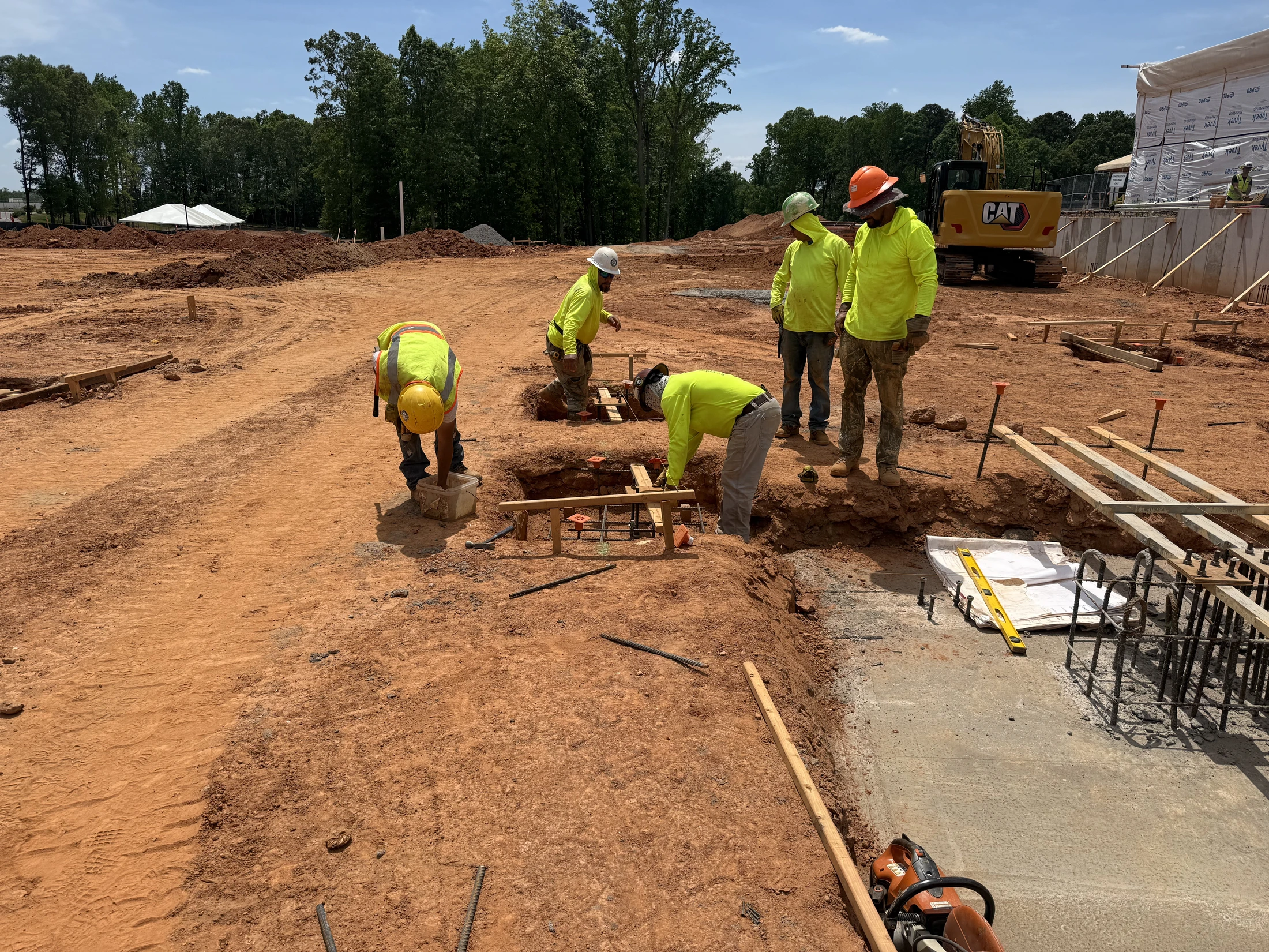 A group of construction workers in hard hats and high-visibility shirts work on setting up rebar for a foundation in a large red dirt construction site.