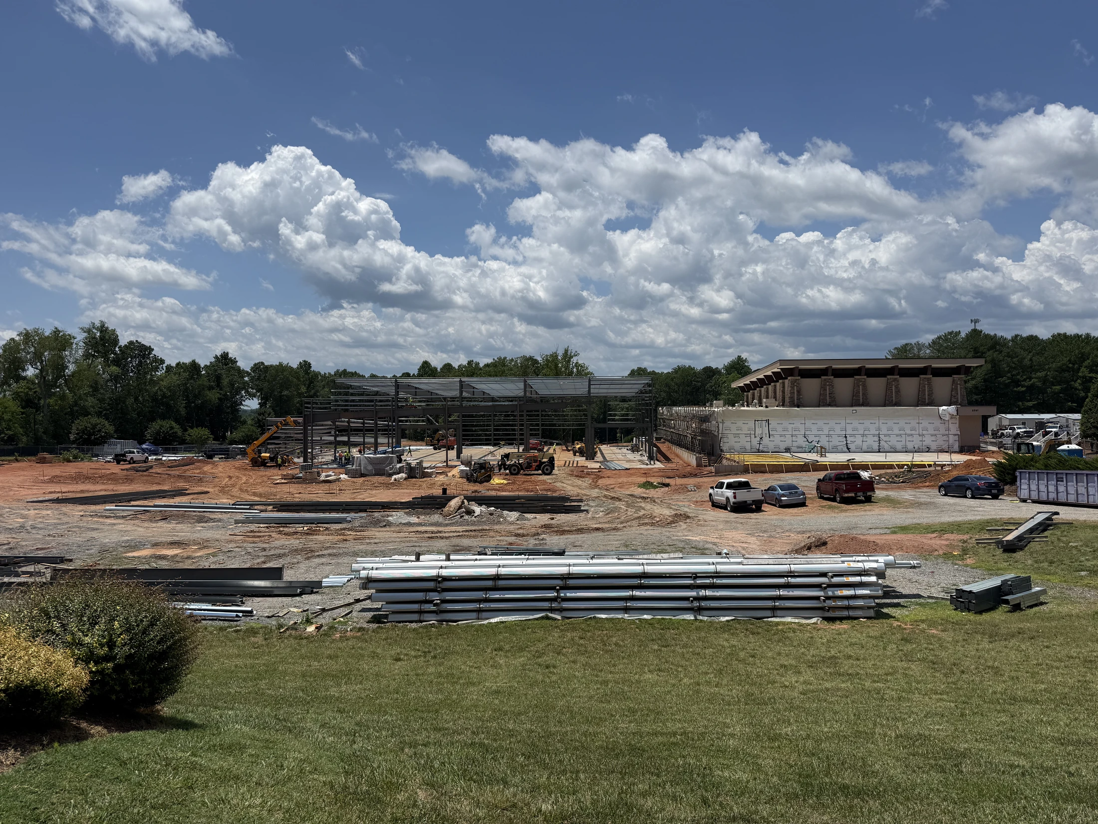 A wide view of a large construction site where the steel frame of a new building is being erected. Materials are stacked on red dirt under a blue, cloudy sky.
