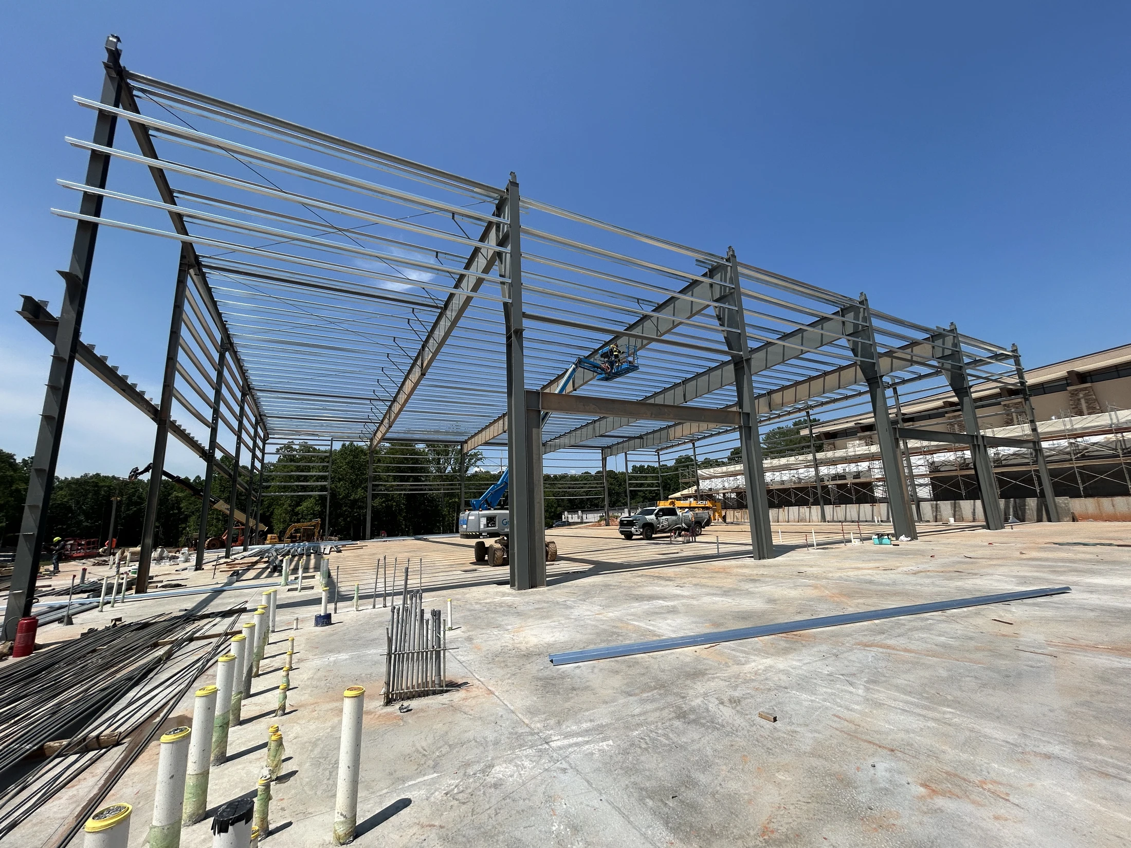 A large steel frame for a new building is being erected on a concrete slab. A worker in an aerial lift is working on the structure under a clear blue sky.