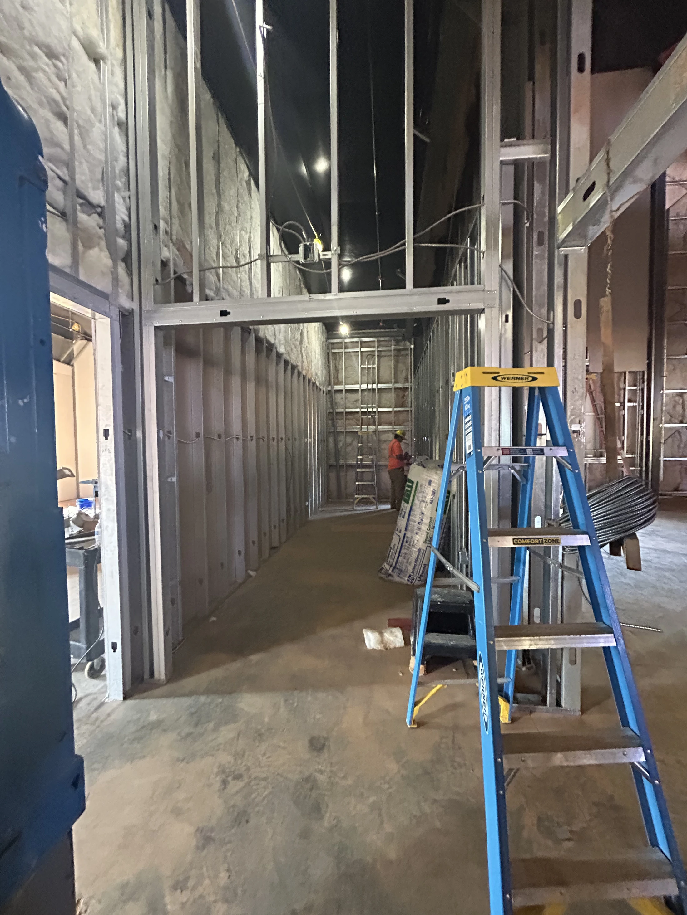 A long hallway in a building under construction, with exposed metal wall studs and insulation. A blue stepladder is prominent in the foreground.