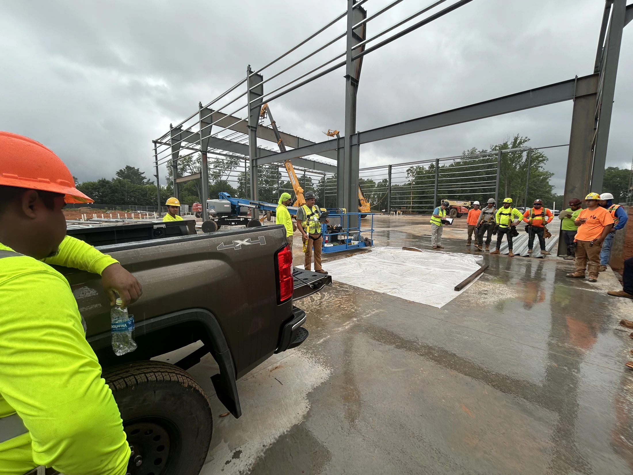 A crew of construction workers in safety gear gathers on a wet concrete slab during the erection of a large steel-frame building on an overcast day.