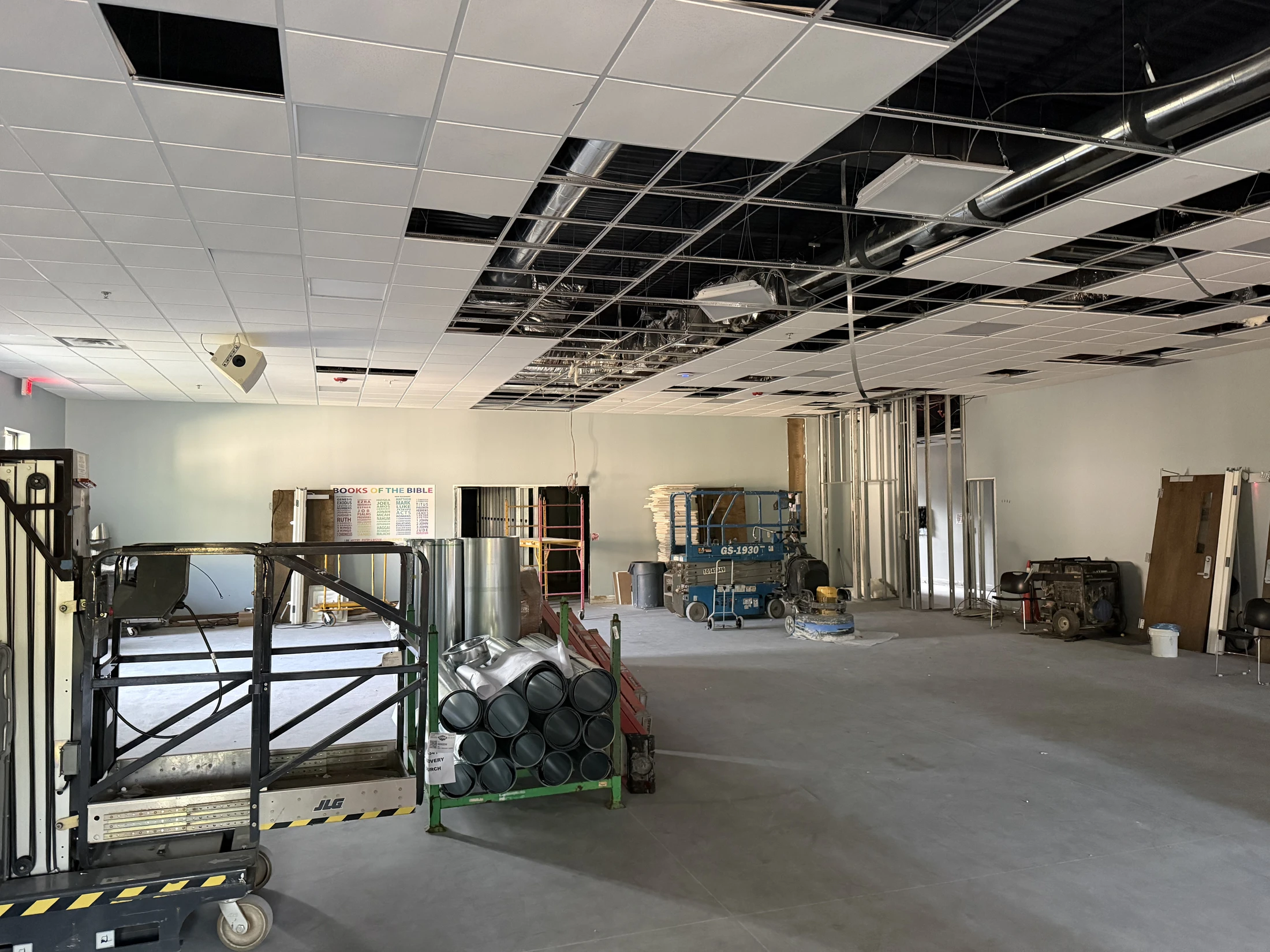 A spacious room during construction, featuring an incomplete drop ceiling with exposed ductwork, concrete floors, and construction lifts and materials.
