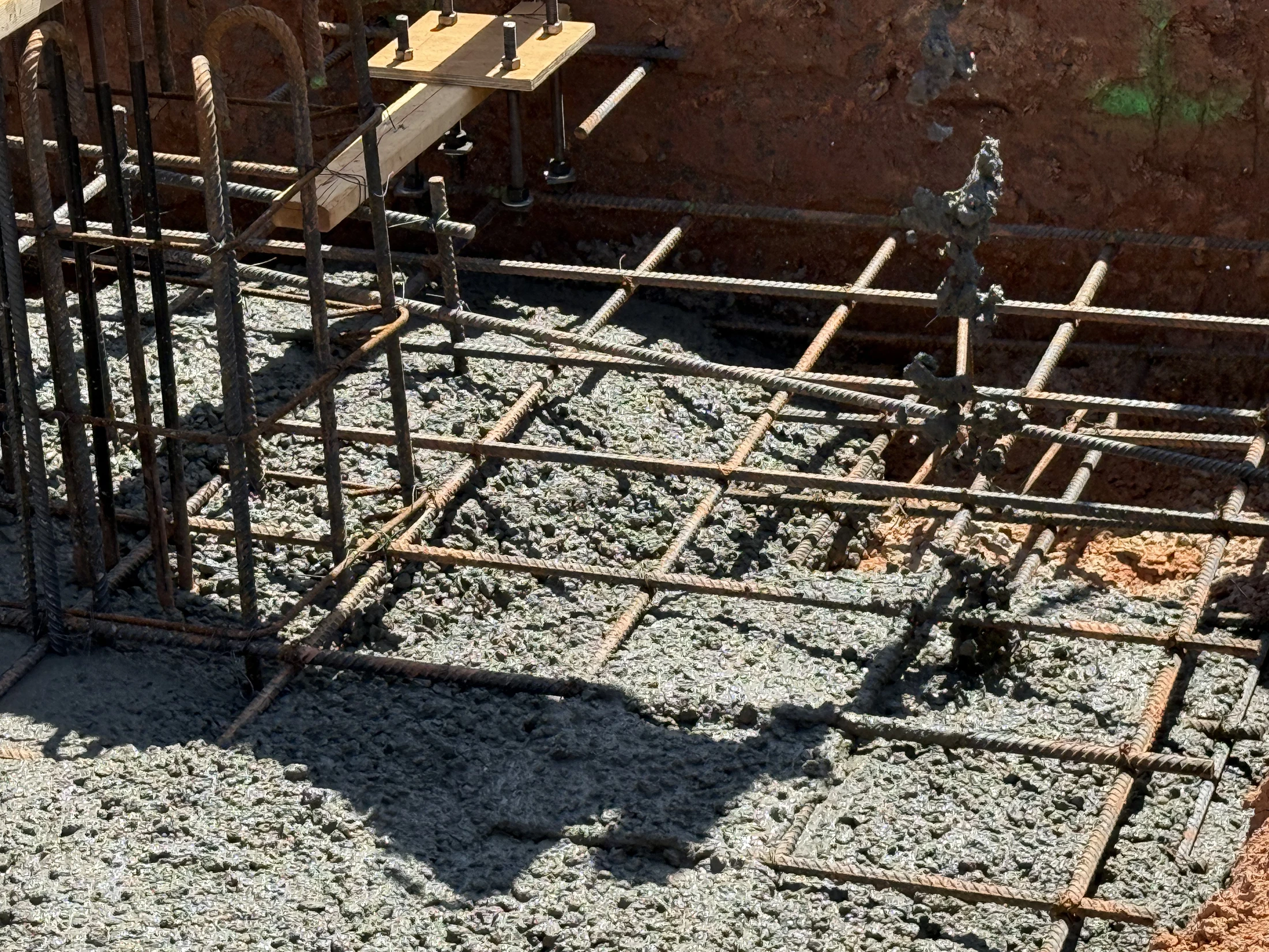 A close-up of a construction site foundation with wet concrete poured around a grid of steel rebar, with anchor bolts set in a wooden form in the background.