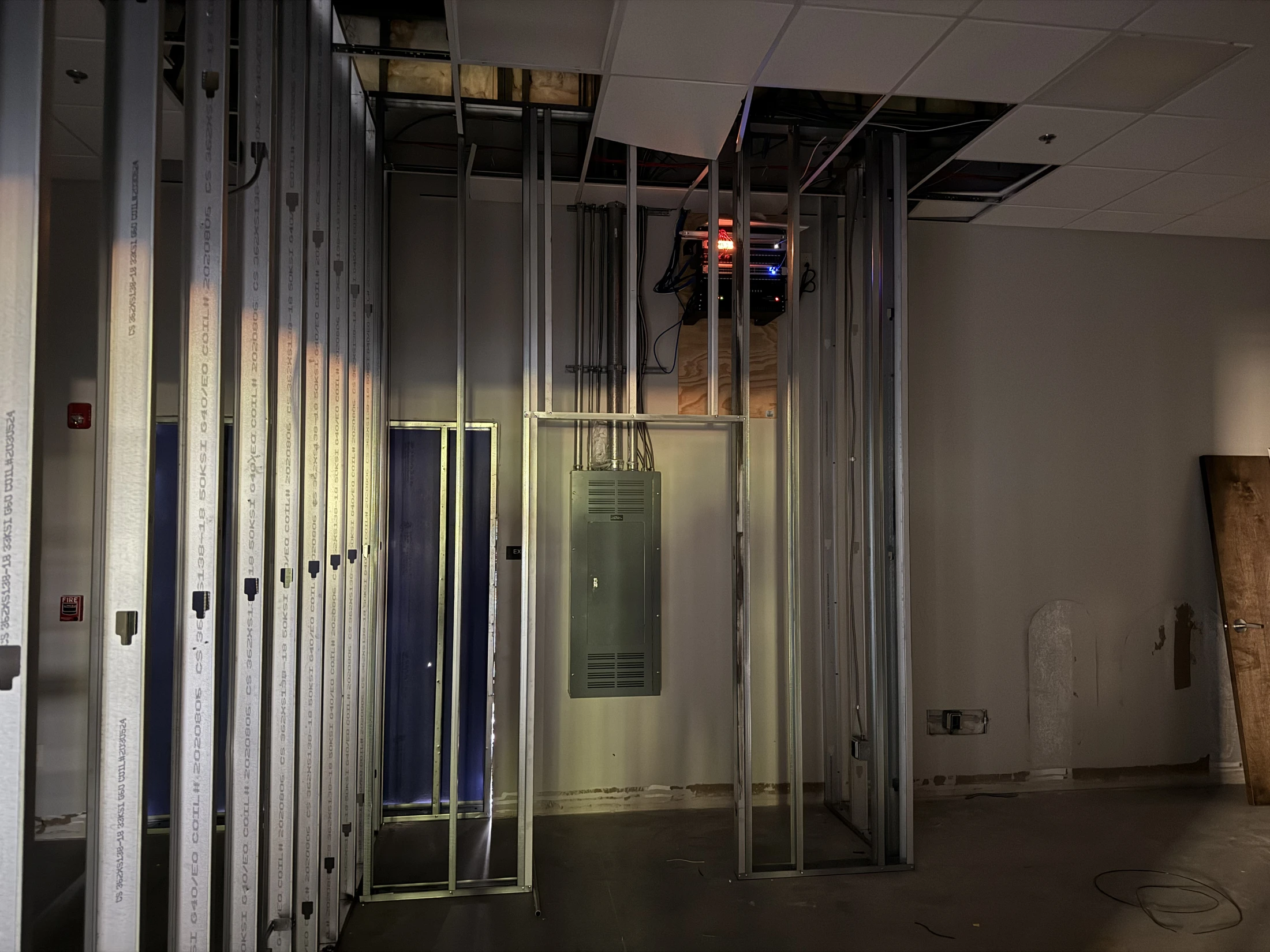 A dimly lit room under construction with exposed metal wall studs, an open ceiling, and an electrical panel and server rack mounted on the far wall.