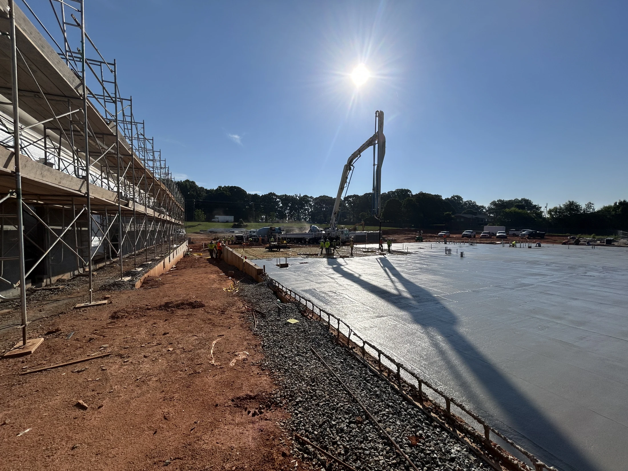 On a bright, sunny day, a concrete pump truck pours a large slab foundation next to a building with scaffolding, as workers smooth the wet concrete.