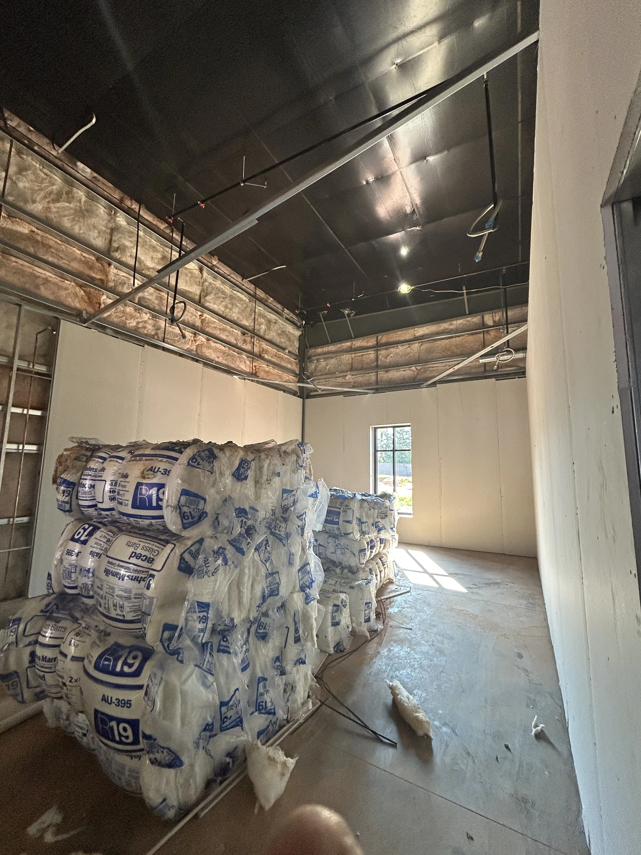 A room under construction with stacks of packaged insulation on the floor. The walls have exposed metal studs and insulation with some drywall installed.