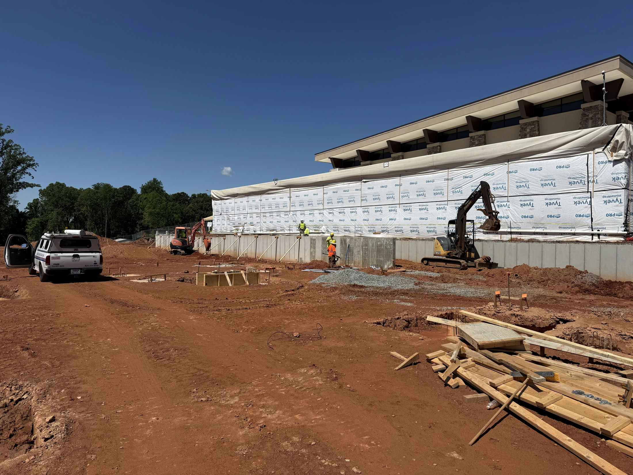 A wide shot of a construction site with a large building foundation, red dirt ground, workers in safety vests, and two excavators under a clear blue sky.