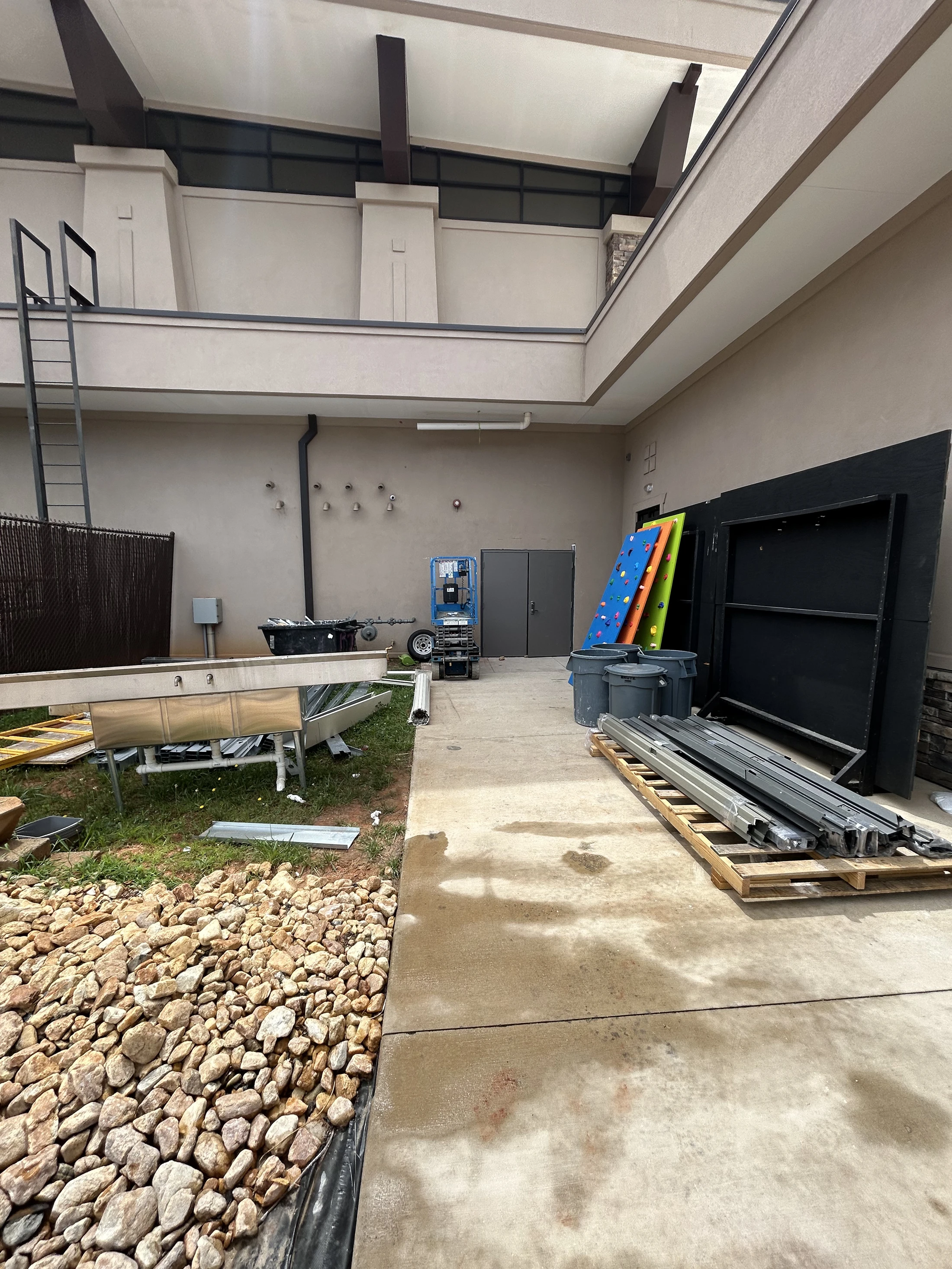 An outdoor area behind a building cluttered with construction materials, including a large metal sink, framing on a pallet, and a scissor lift.