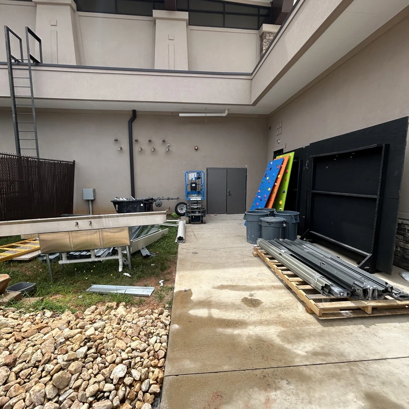 An outdoor area behind a building cluttered with construction materials, including a large metal sink, framing on a pallet, and a scissor lift.