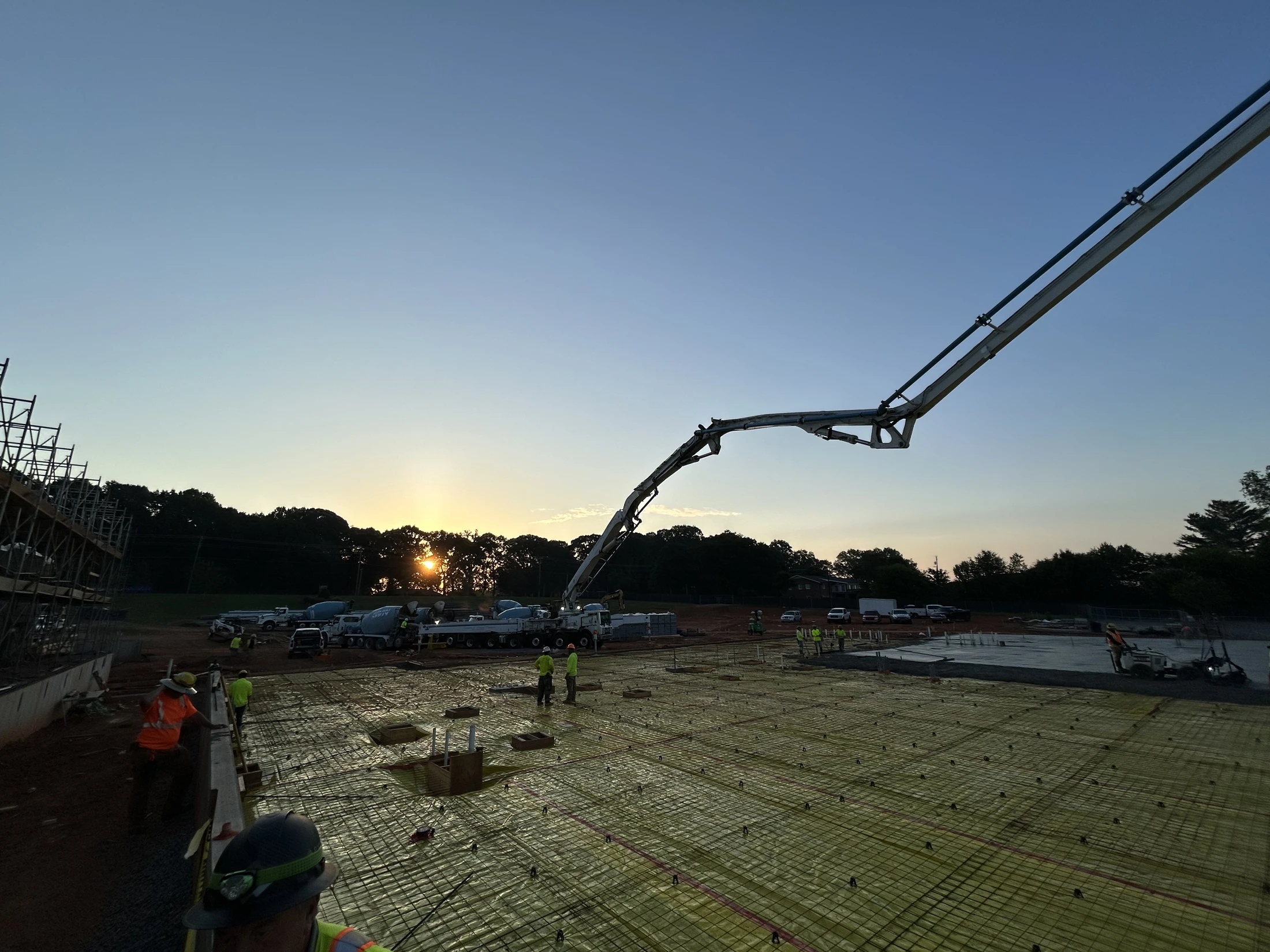 A concrete pump truck extends its boom over a large rebar grid foundation at a construction site during sunrise, with workers and mixer trucks nearby.