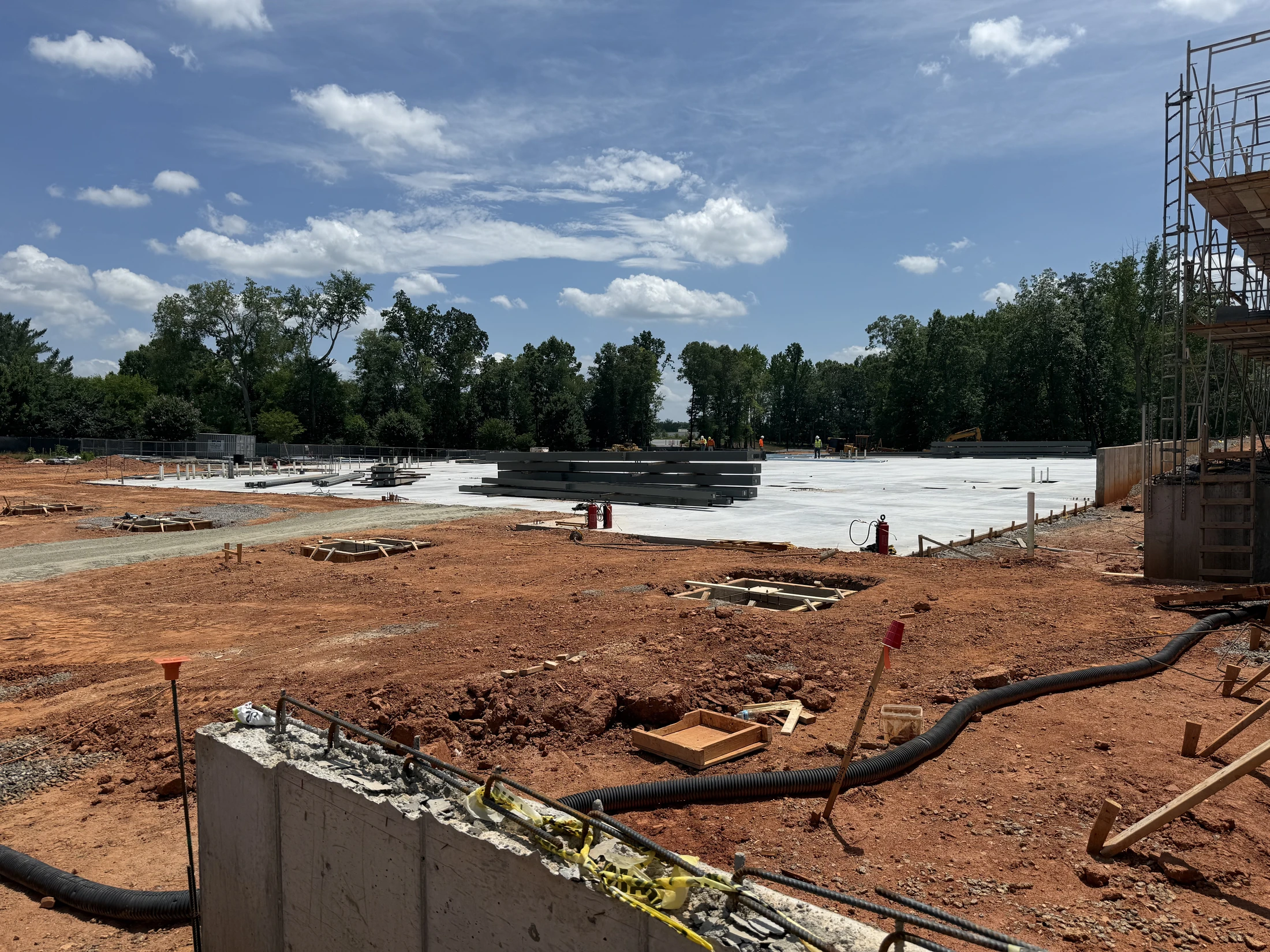 A large construction site with red clay ground, a newly poured concrete foundation, and stacked steel beams under a partly cloudy blue sky.