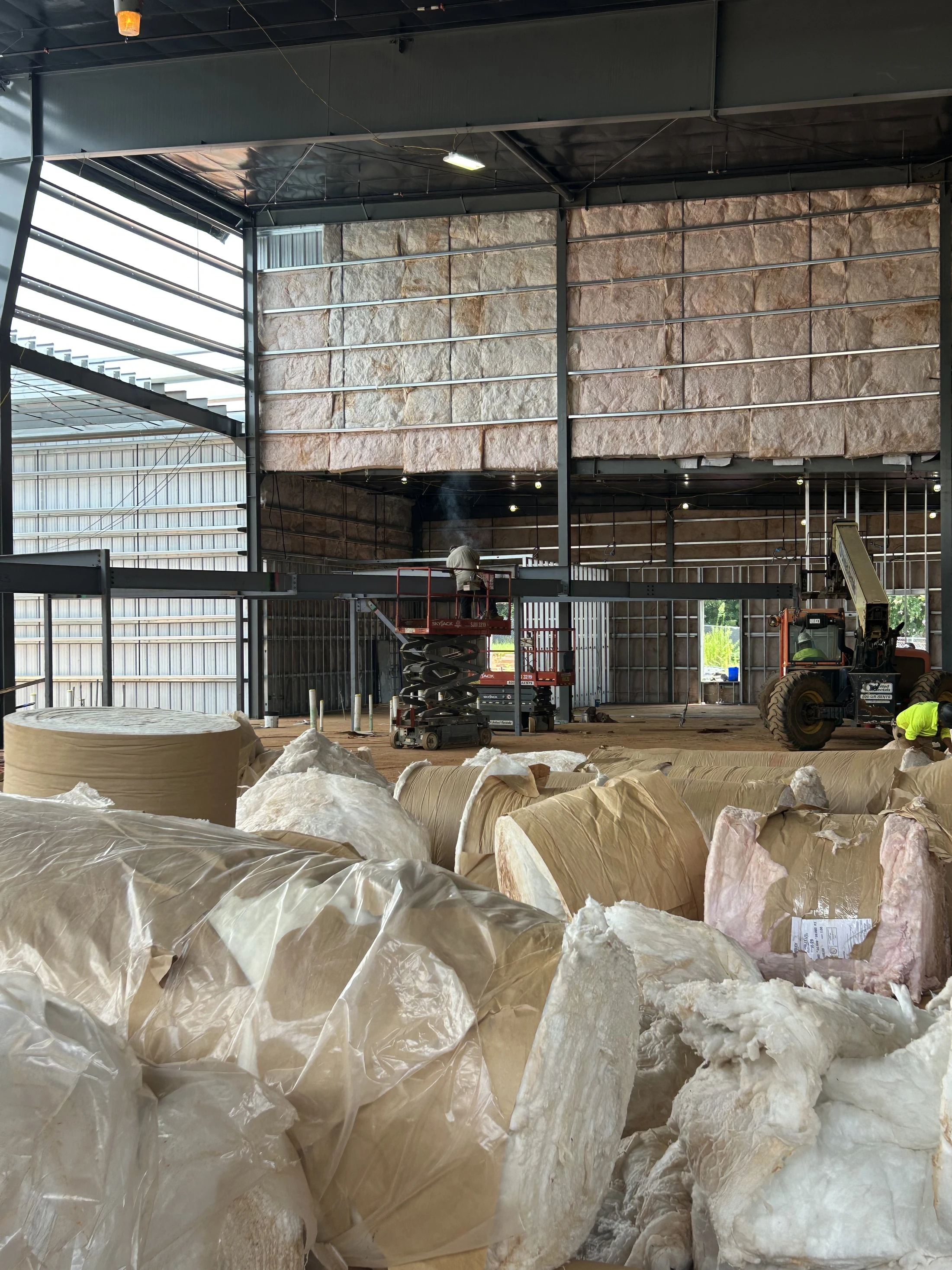 The interior of a large steel-frame building under construction, with workers installing insulation into the walls and large rolls of material on the ground.