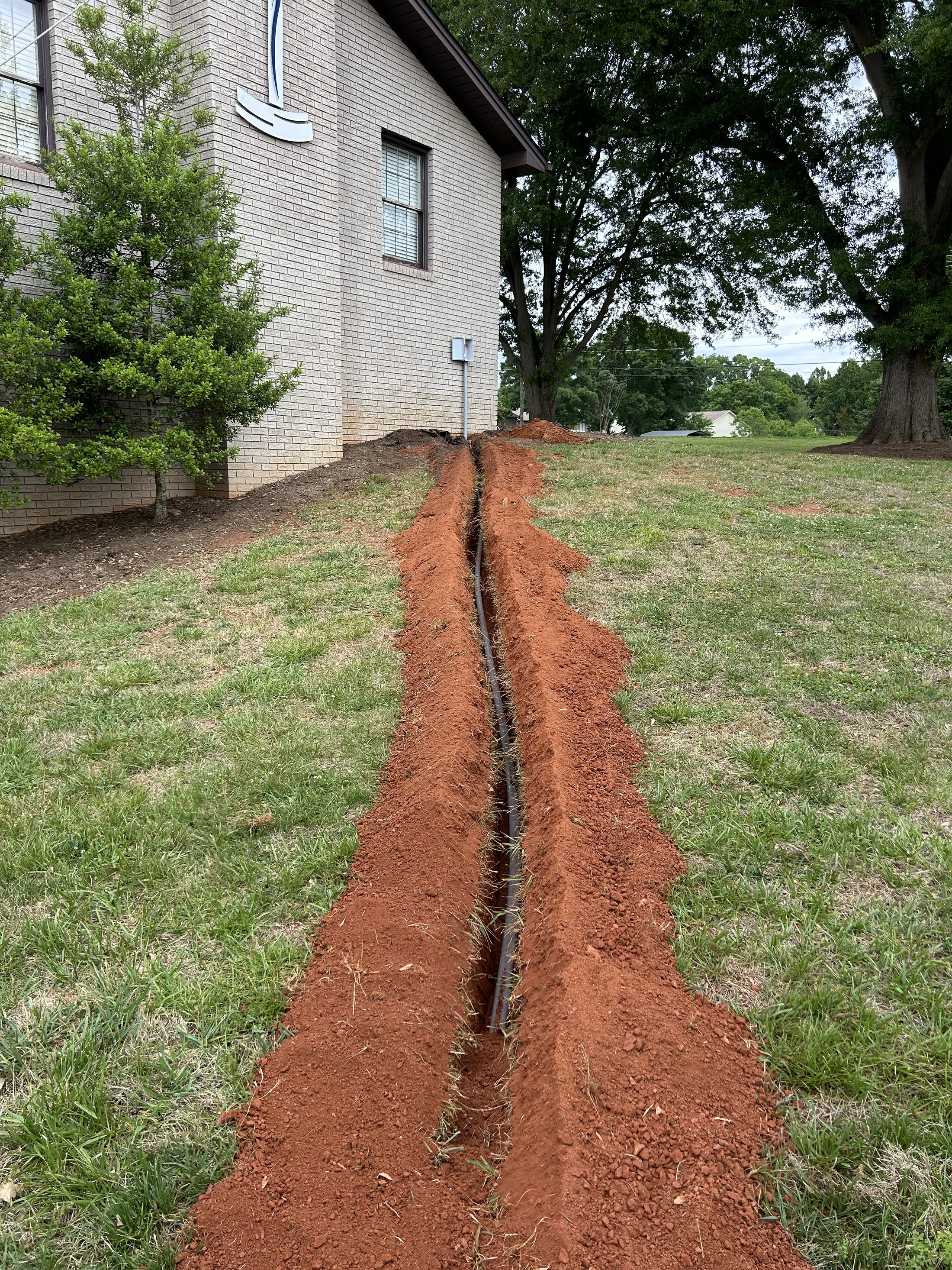 A long, narrow trench dug in a grassy yard next to a brick building, with a black pipe laid inside and reddish-brown excavated soil piled on either side.