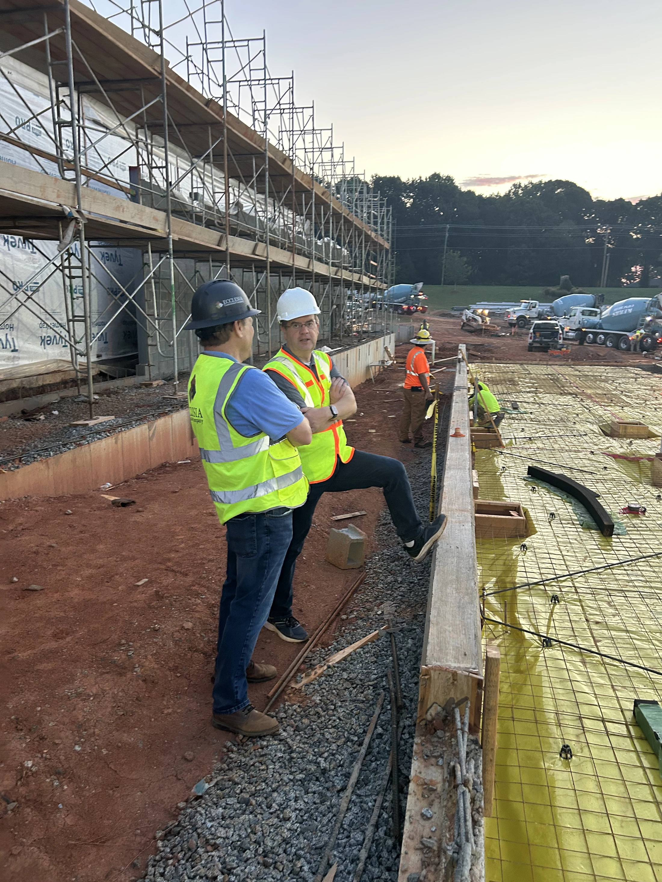 Two men in hard hats and safety vests talk on a construction site next to a foundation being prepared for a concrete pour, with more workers in the background.