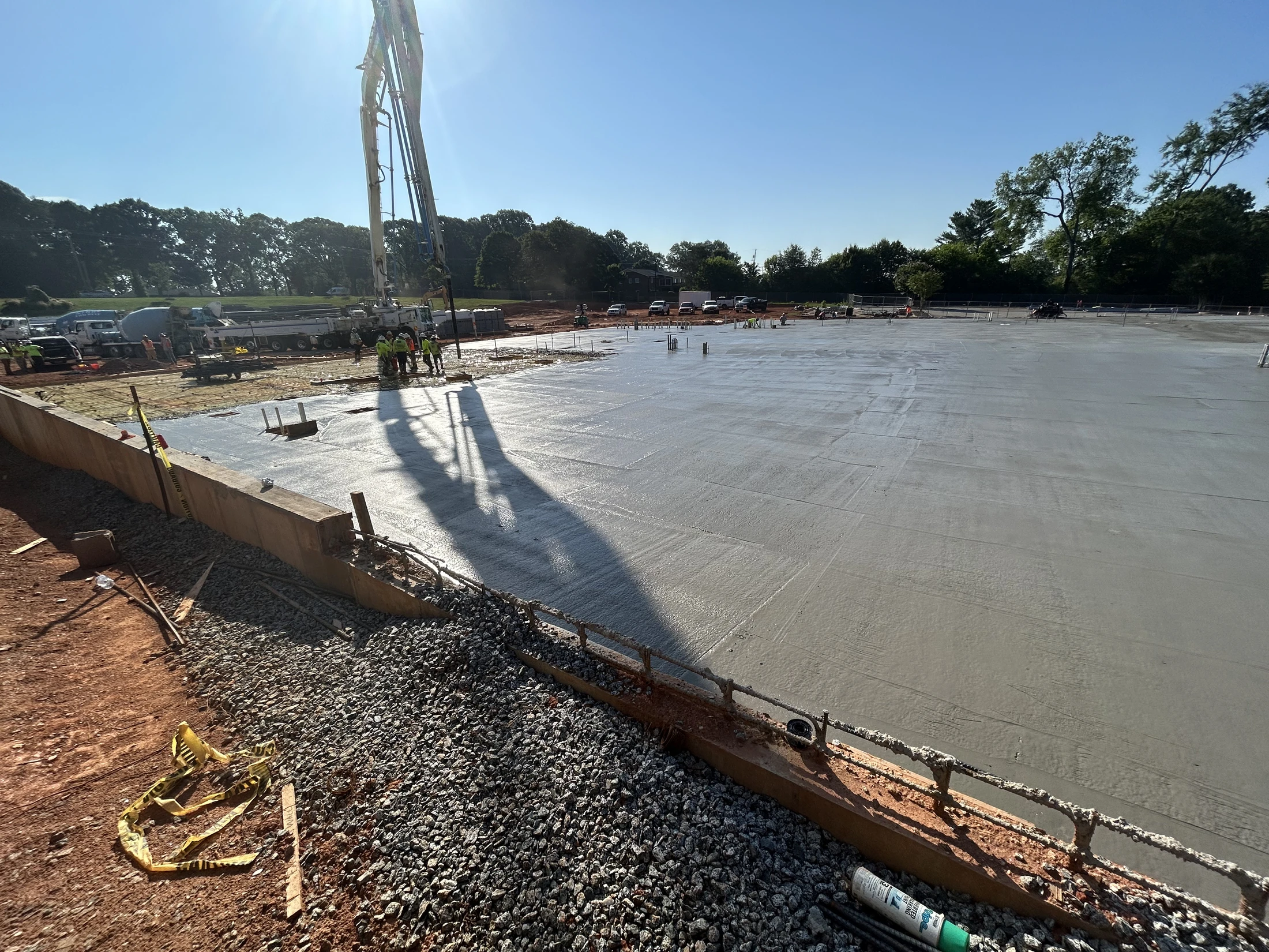 A large concrete slab is being poured by a pump truck on a sunny construction site. The pump's long shadow stretches across the wet, grey surface.