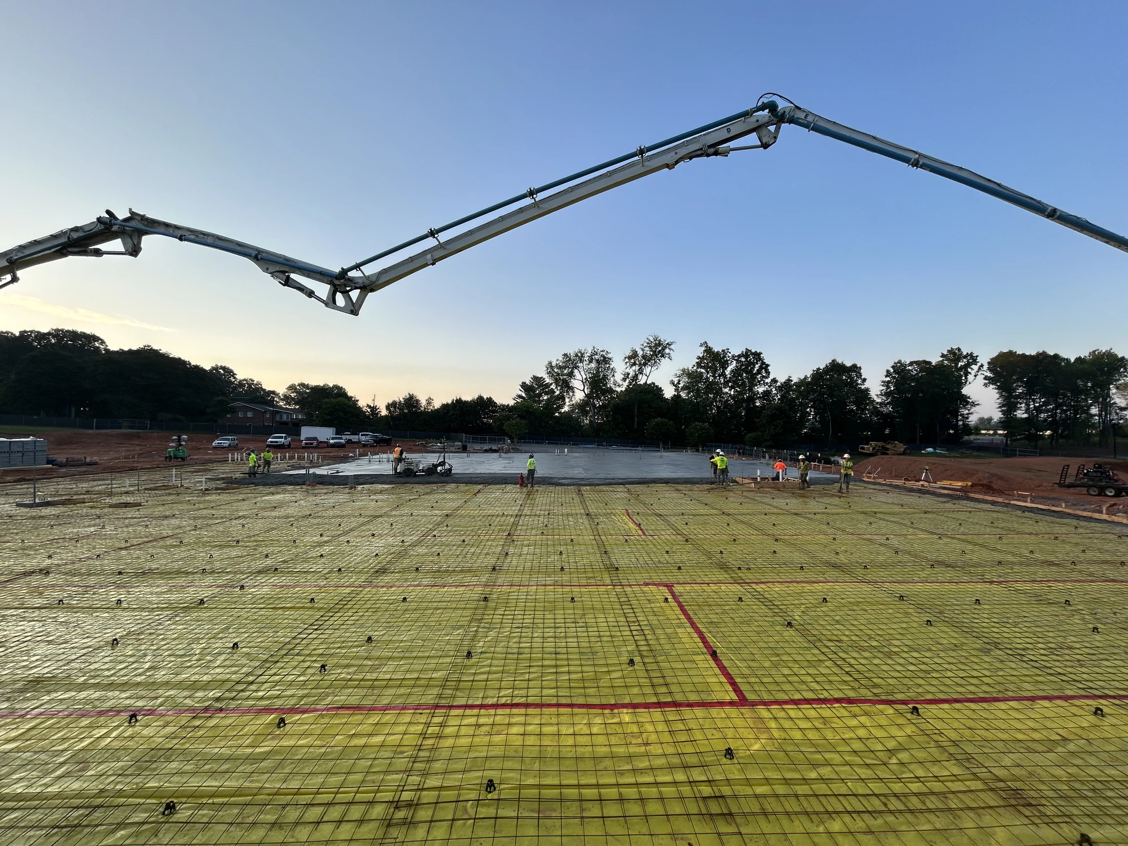 A concrete pump truck's boom extends over a large construction area prepared with rebar, as workers in the background pour a concrete slab at dawn.