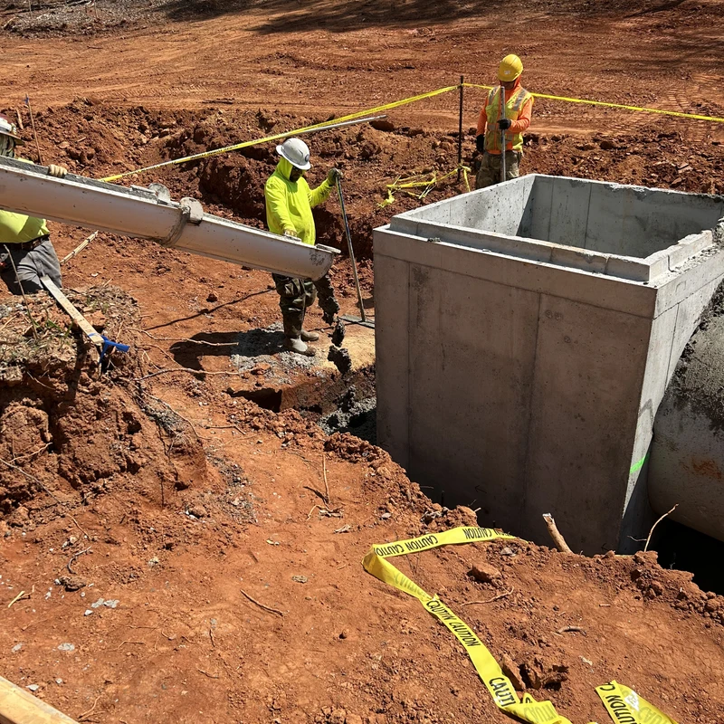 Three construction workers in safety gear pour wet concrete from a chute into the ground next to a large concrete box on a red clay construction site.