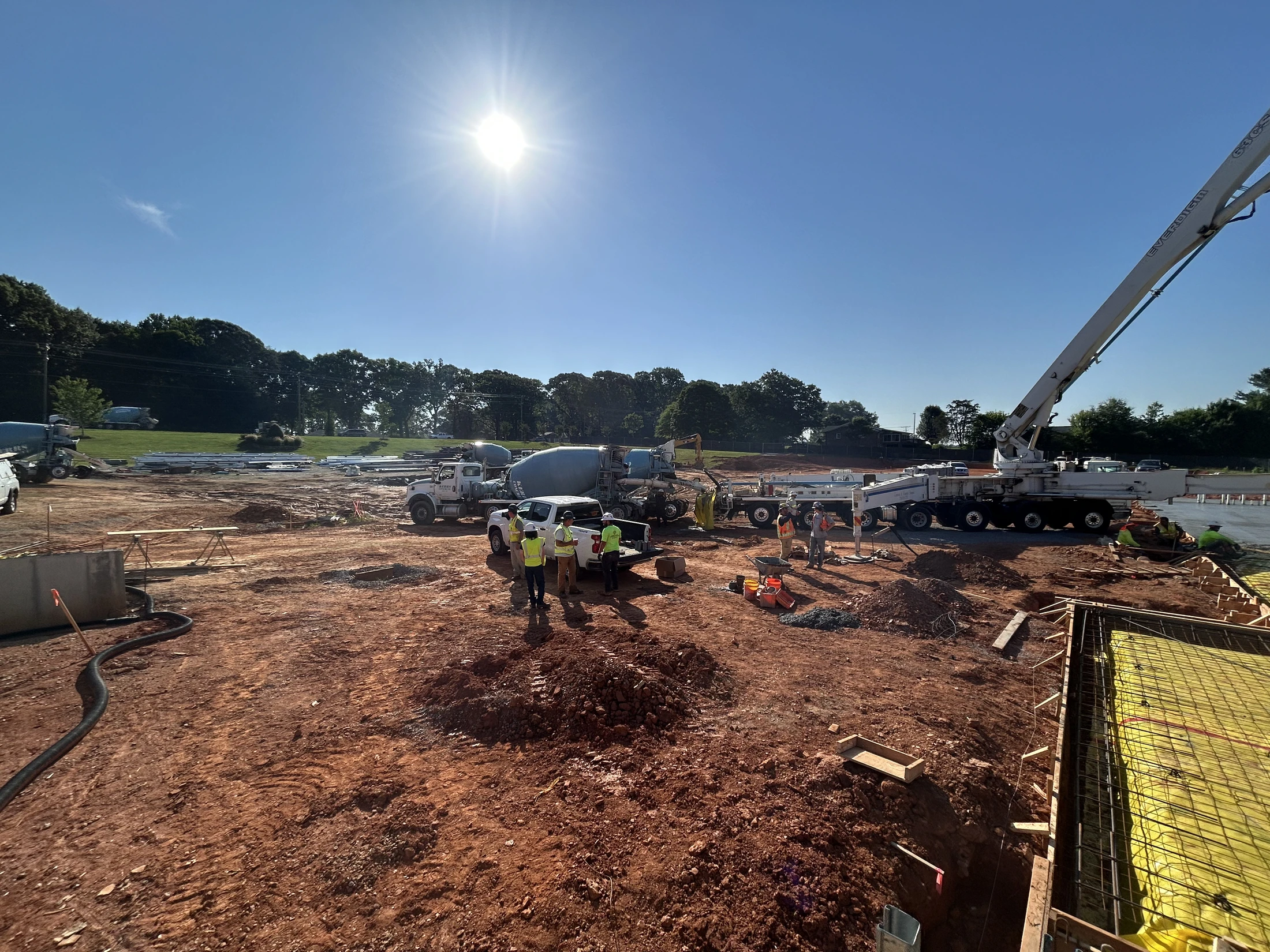 A wide shot of a construction site under a bright sun. Workers gather around a concrete mixer and a large pump truck on a red dirt lot next to a foundation.