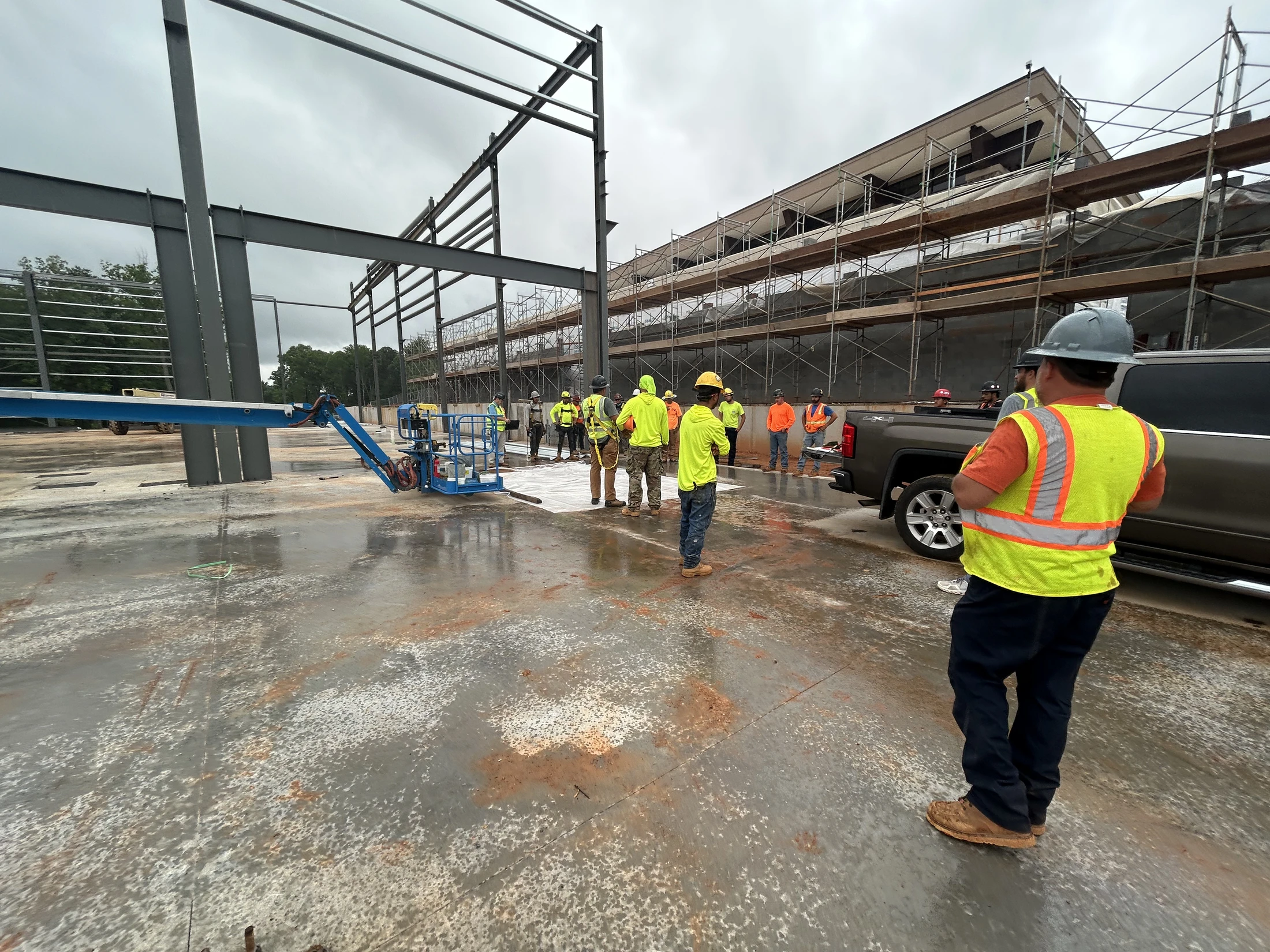 A group of construction workers in hard hats and safety vests stand on a wet concrete slab at a building site with a new steel frame structure in progress.