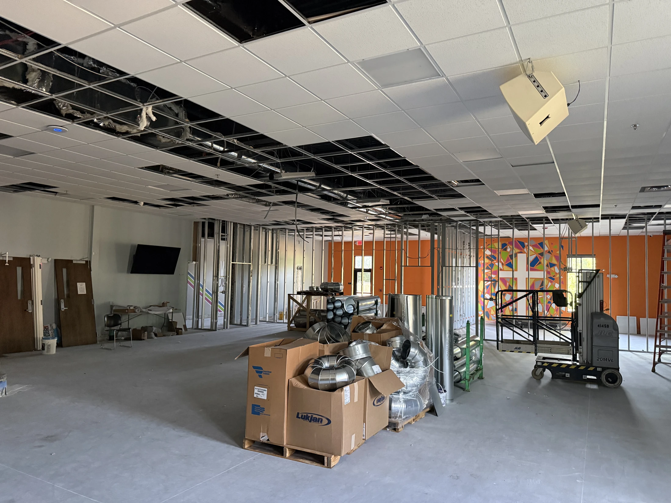 A large room under construction with metal wall framing, an incomplete drop ceiling, and boxes of ductwork on a pallet in the center of the concrete floor.