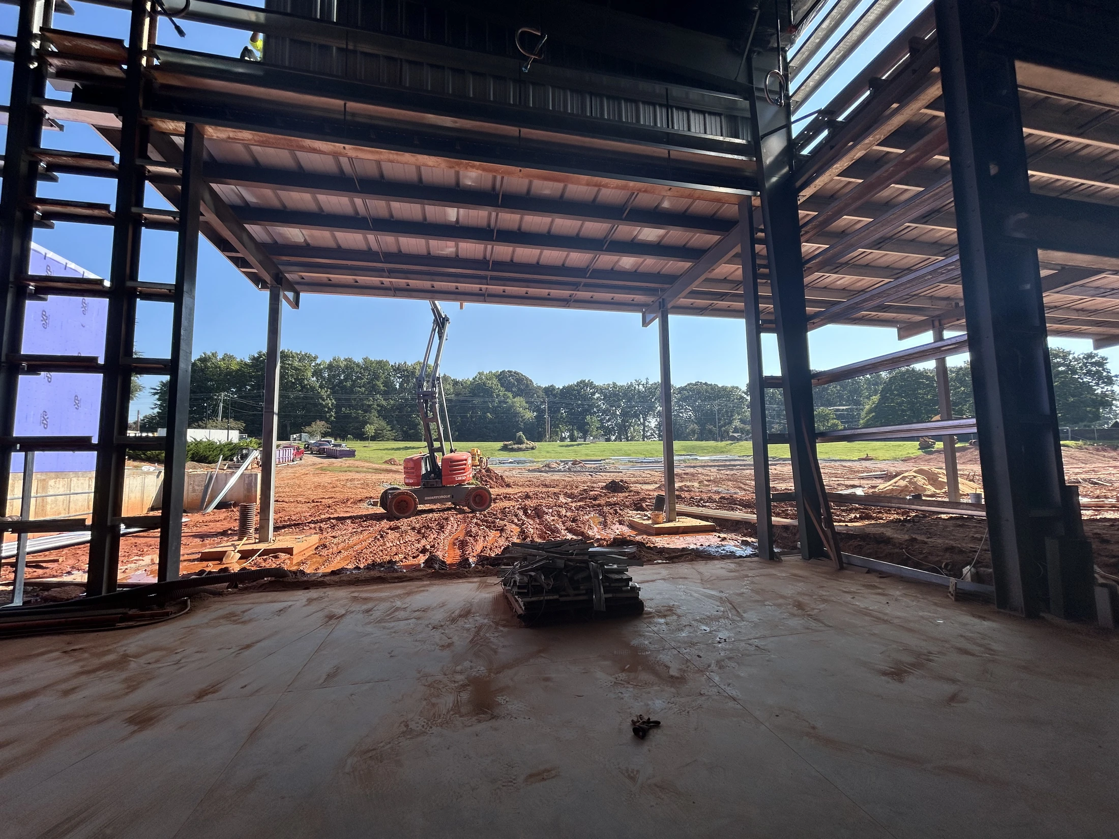View from inside a steel-framed building looking out onto a muddy construction site with heavy machinery. A green field and trees are in the background.