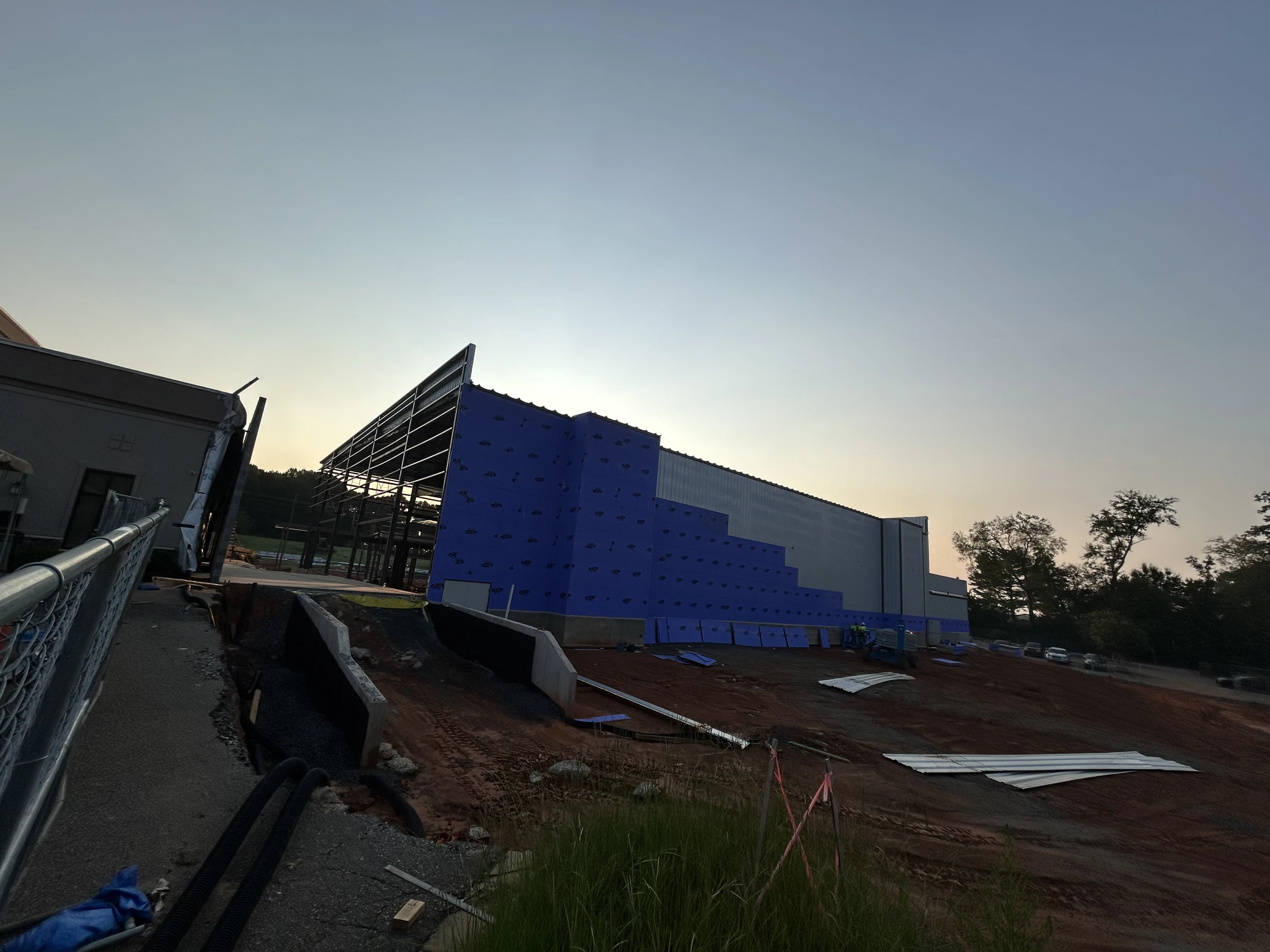 A wide shot of a large building on a construction site at dusk. The building has an exposed steel frame with blue and gray exterior walls being installed.