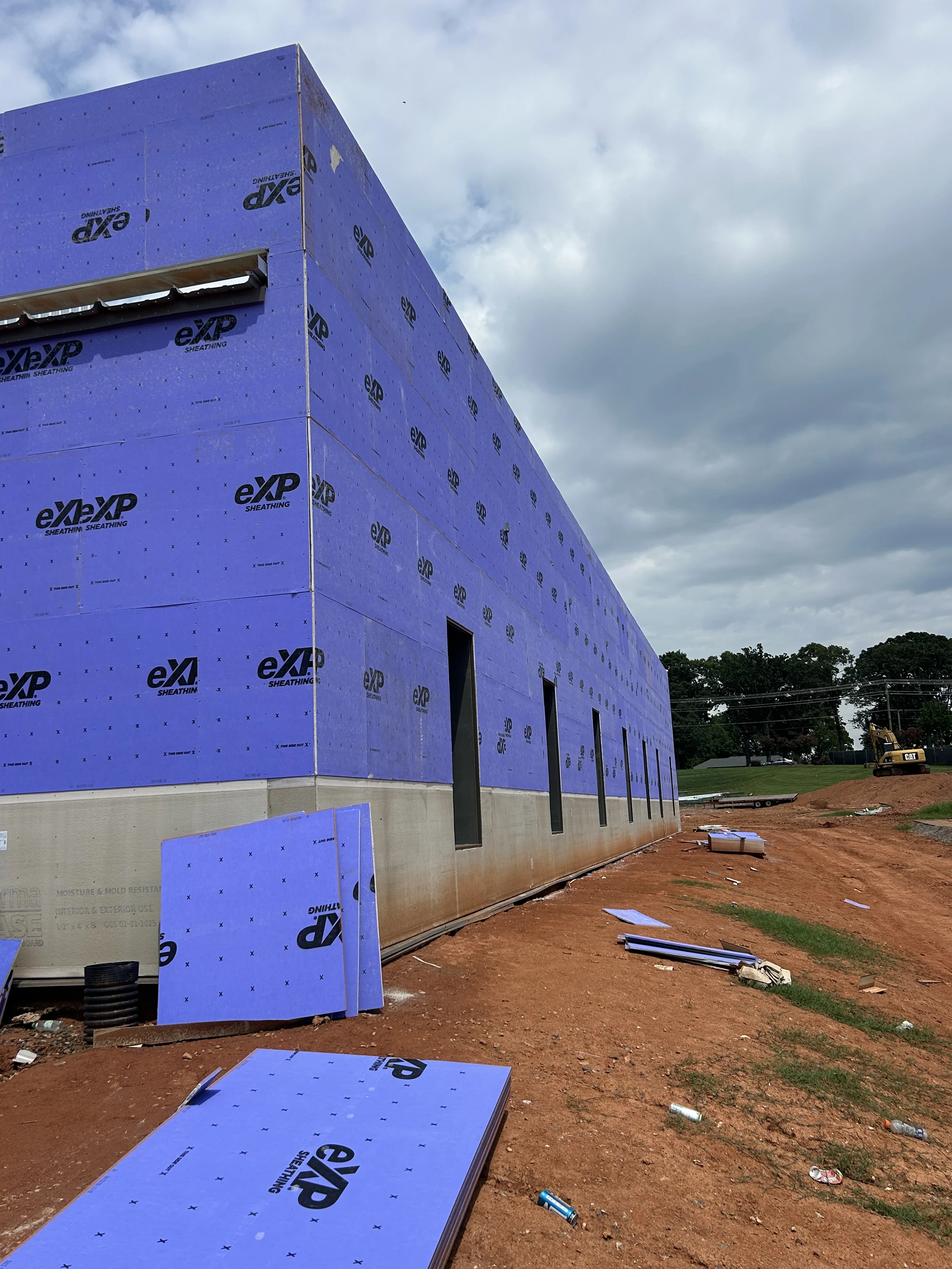 A long commercial building under construction, its exterior walls covered in bright purple sheathing panels. The site is dirt with materials scattered around.