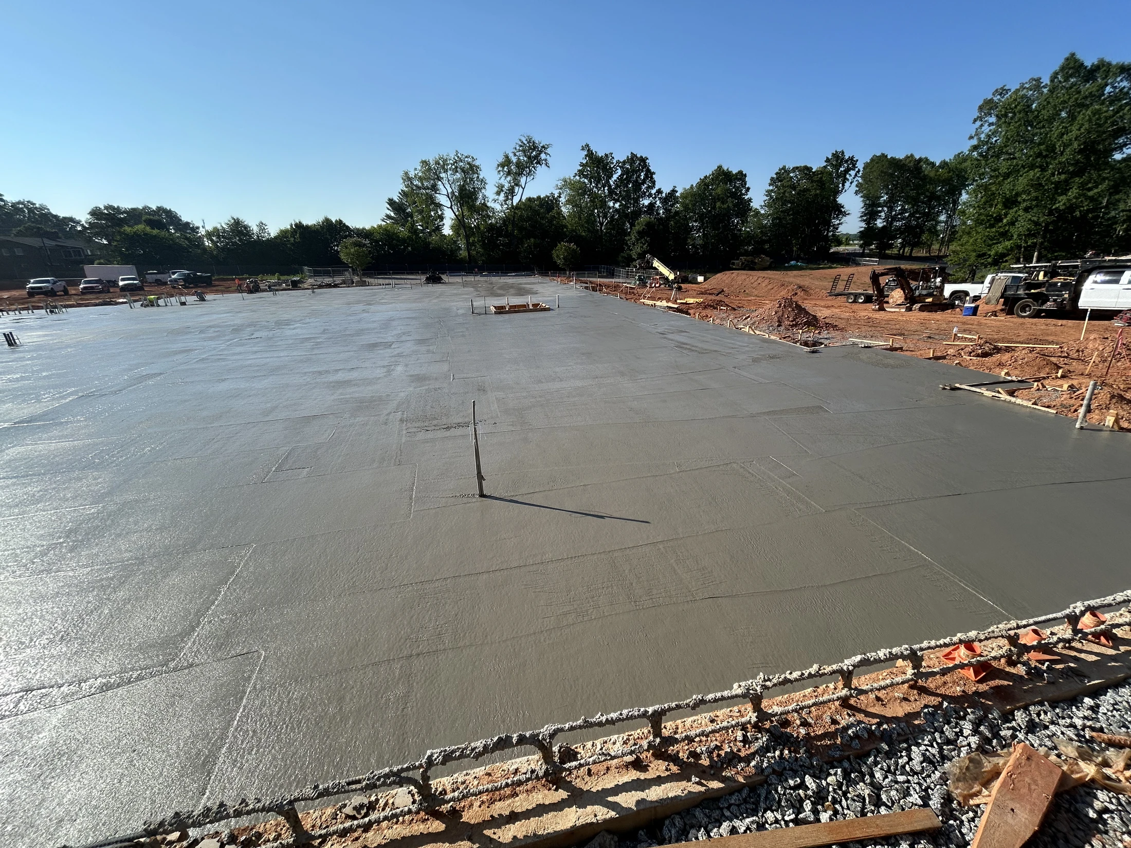 A wide expanse of a freshly poured, wet concrete slab at a construction site on a sunny day. In the background are trees and construction vehicles.