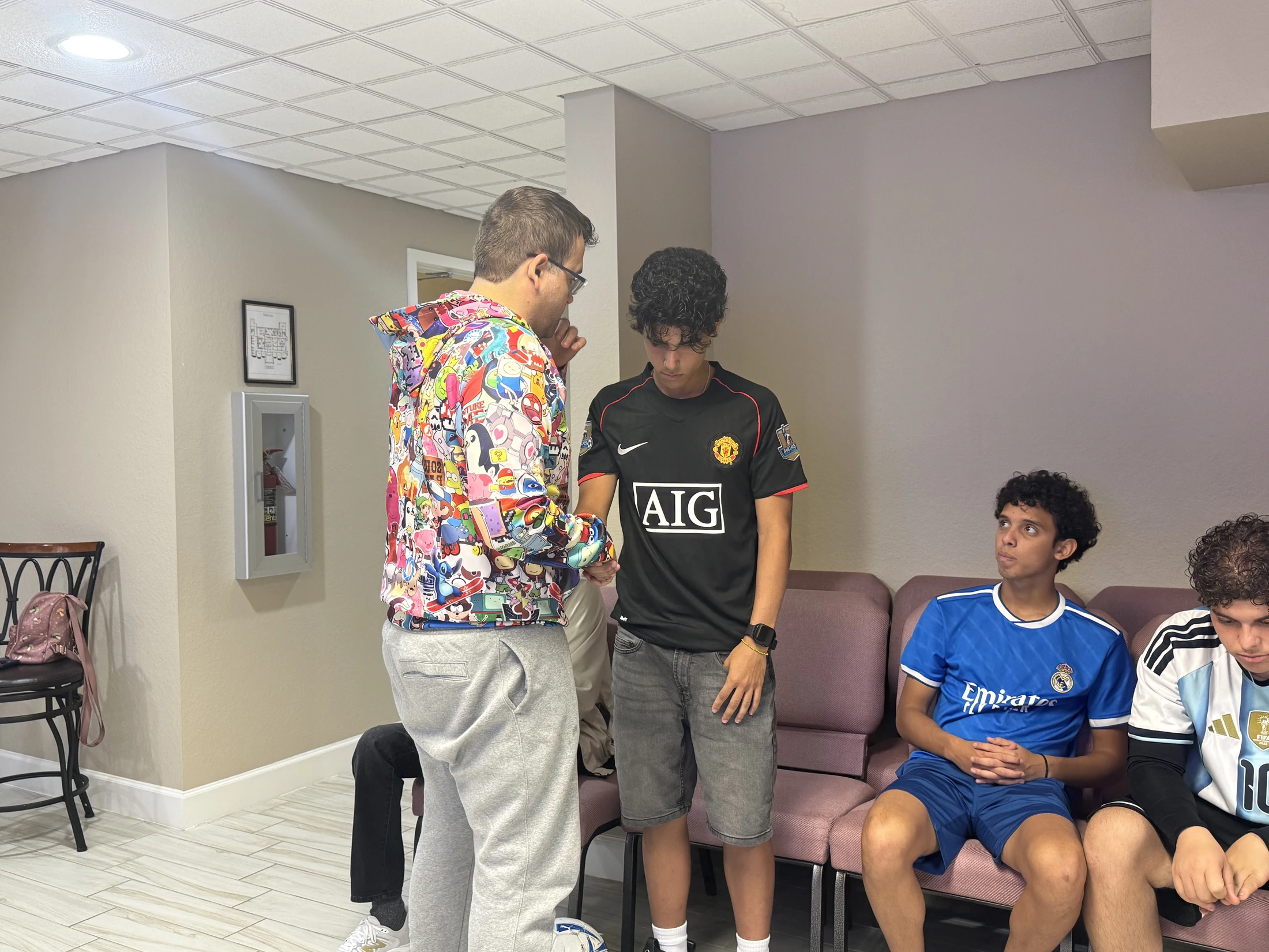 A man in a colorful cartoon-print hoodie speaks to a young man in a soccer jersey, while two other young men in jerseys sit and watch from a bench.