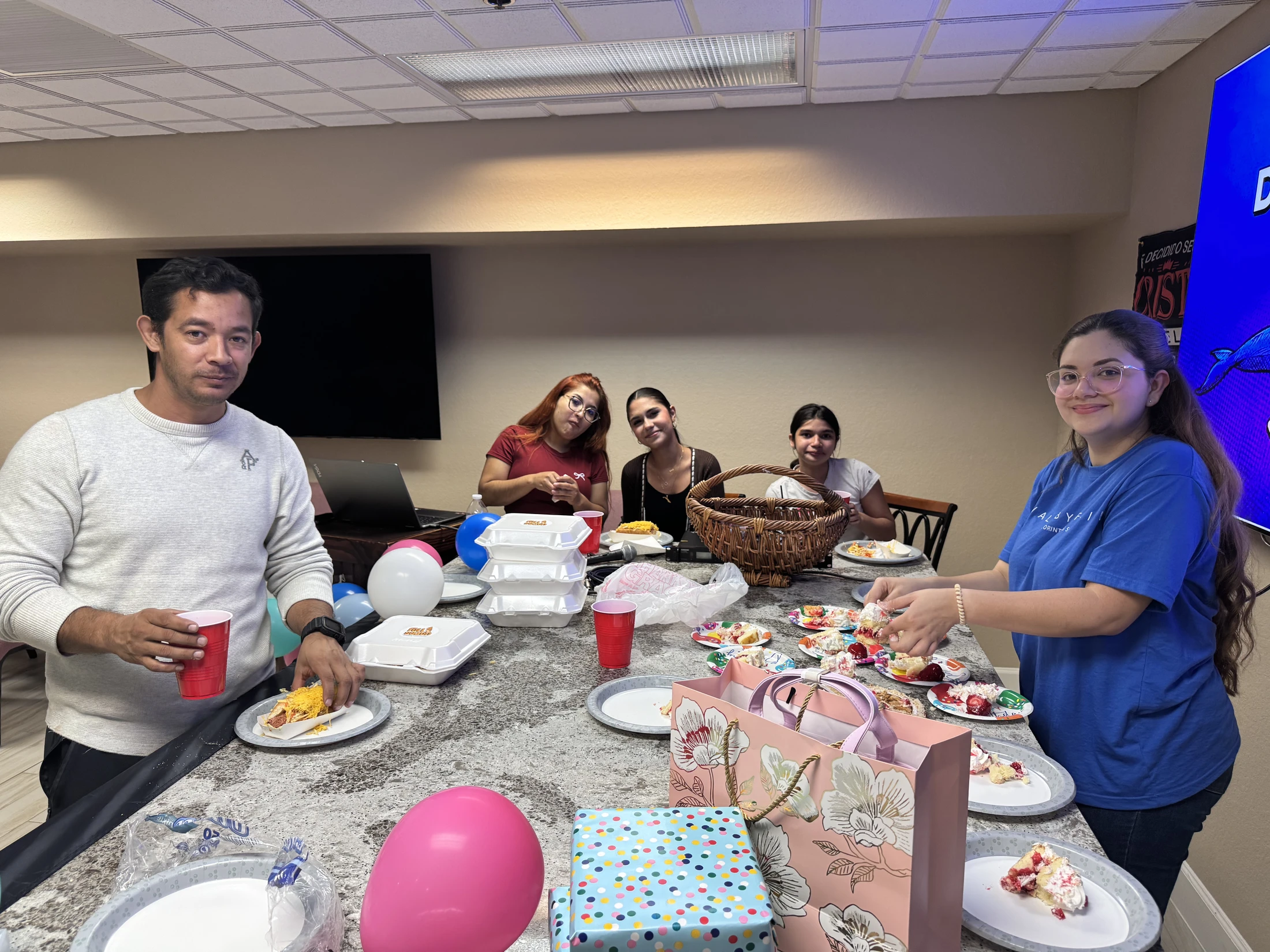 A group of five people celebrating indoors around a counter with food, cake, presents, and balloons, smiling for the camera.