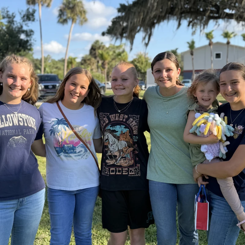 A group of six smiling girls and young women of varying ages stand together on a grassy lawn with palm trees in the background on a sunny day.