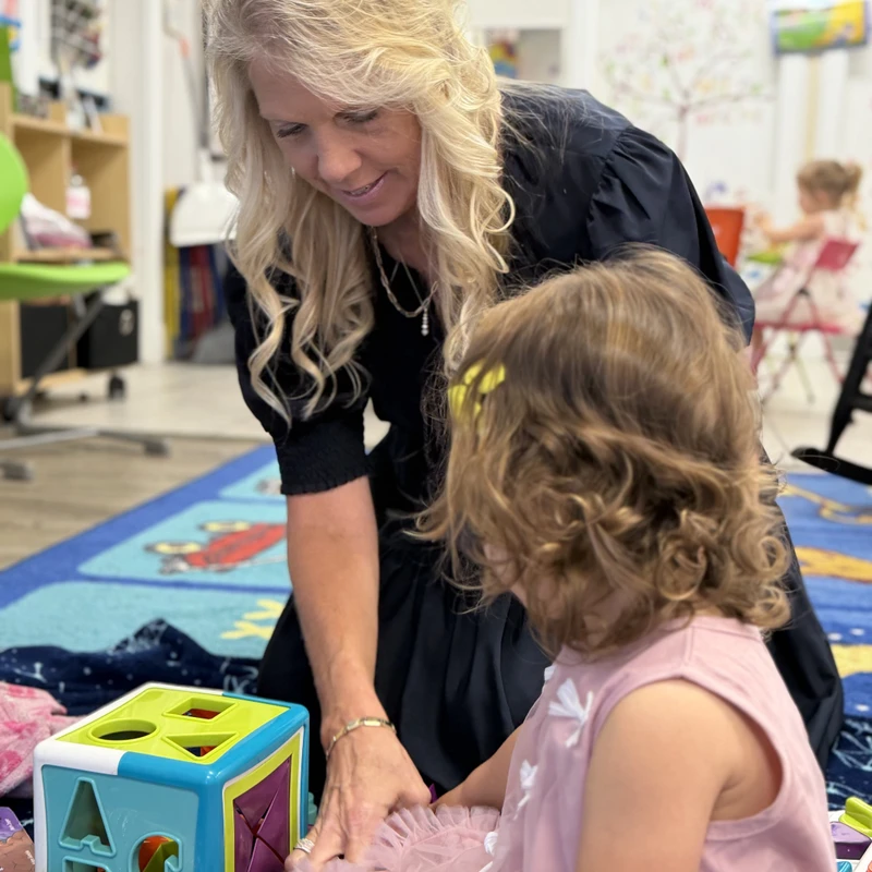 A female teacher with blonde hair kneels on the floor, smiling as she plays with a young girl in a pink dress and a colorful shape-sorter toy.